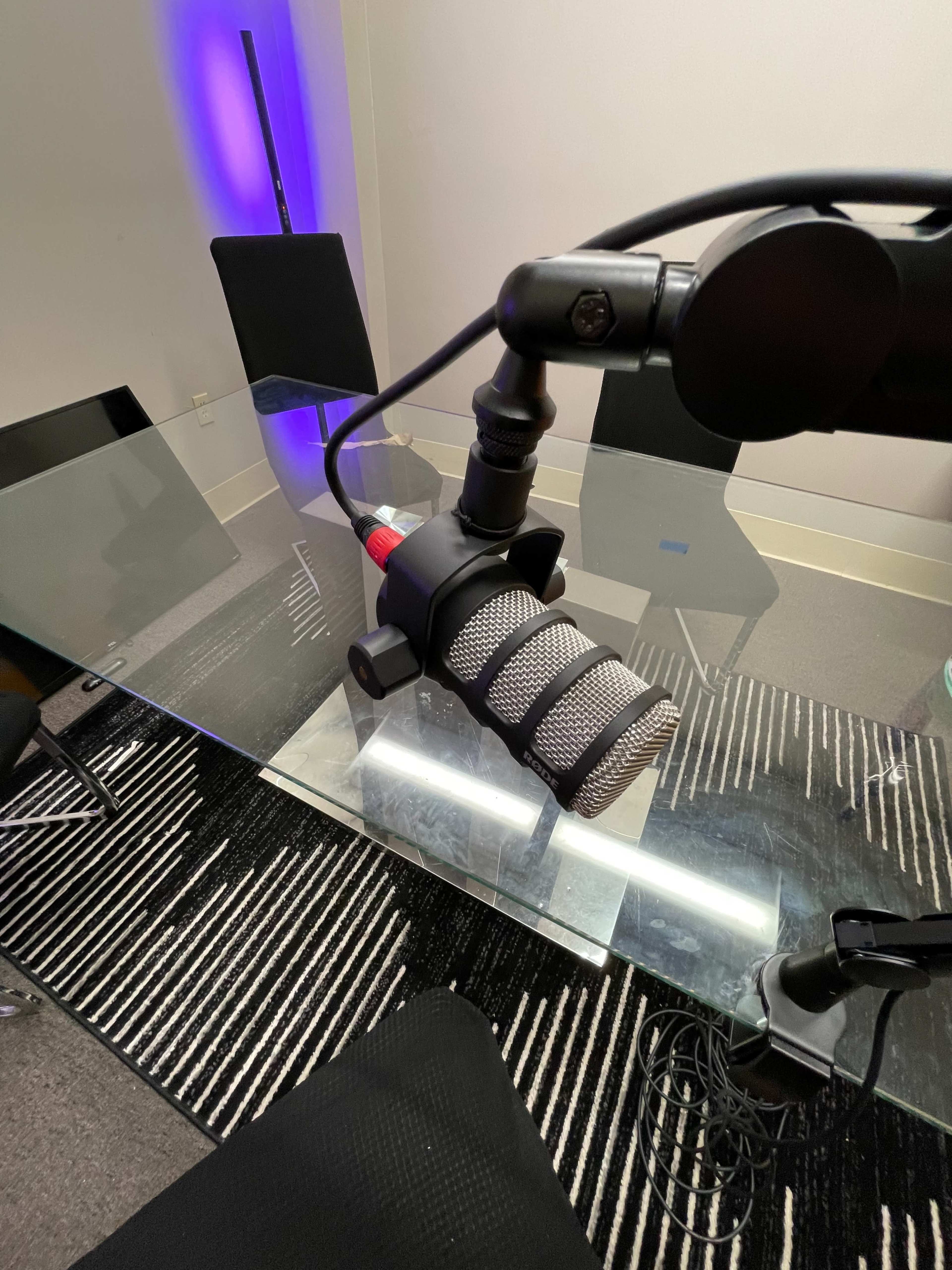 A microphone is positioned on a glass table with a striped rug underneath, surrounded by empty chairs and a purple light in the background.