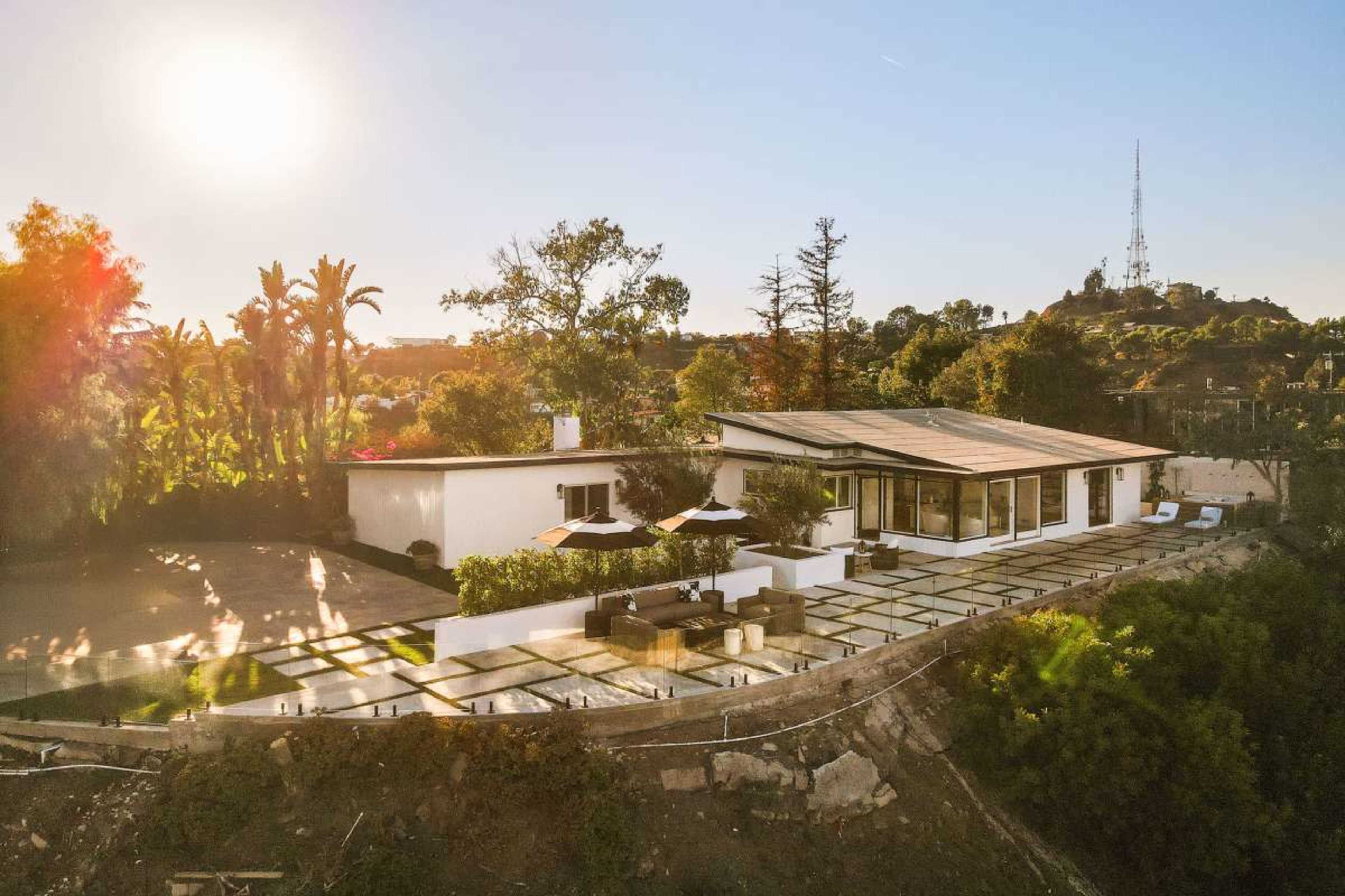 A modern house sits on a hillside with a large patio area surrounded by trees and plants, under a clear blue sky.