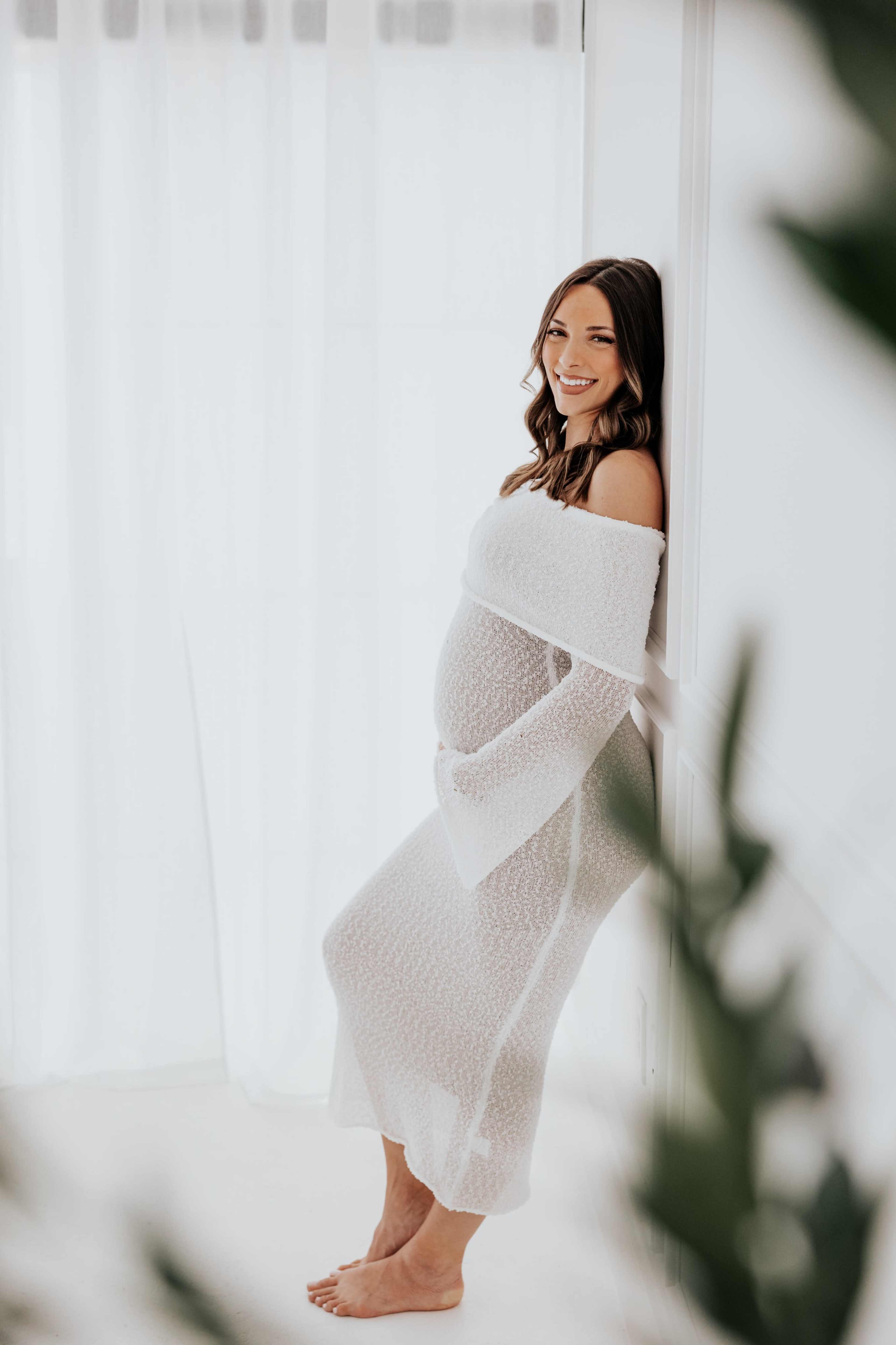 A pregnant woman in a white, off-the-shoulder dress leans against a wall in a bright, softly lit room.