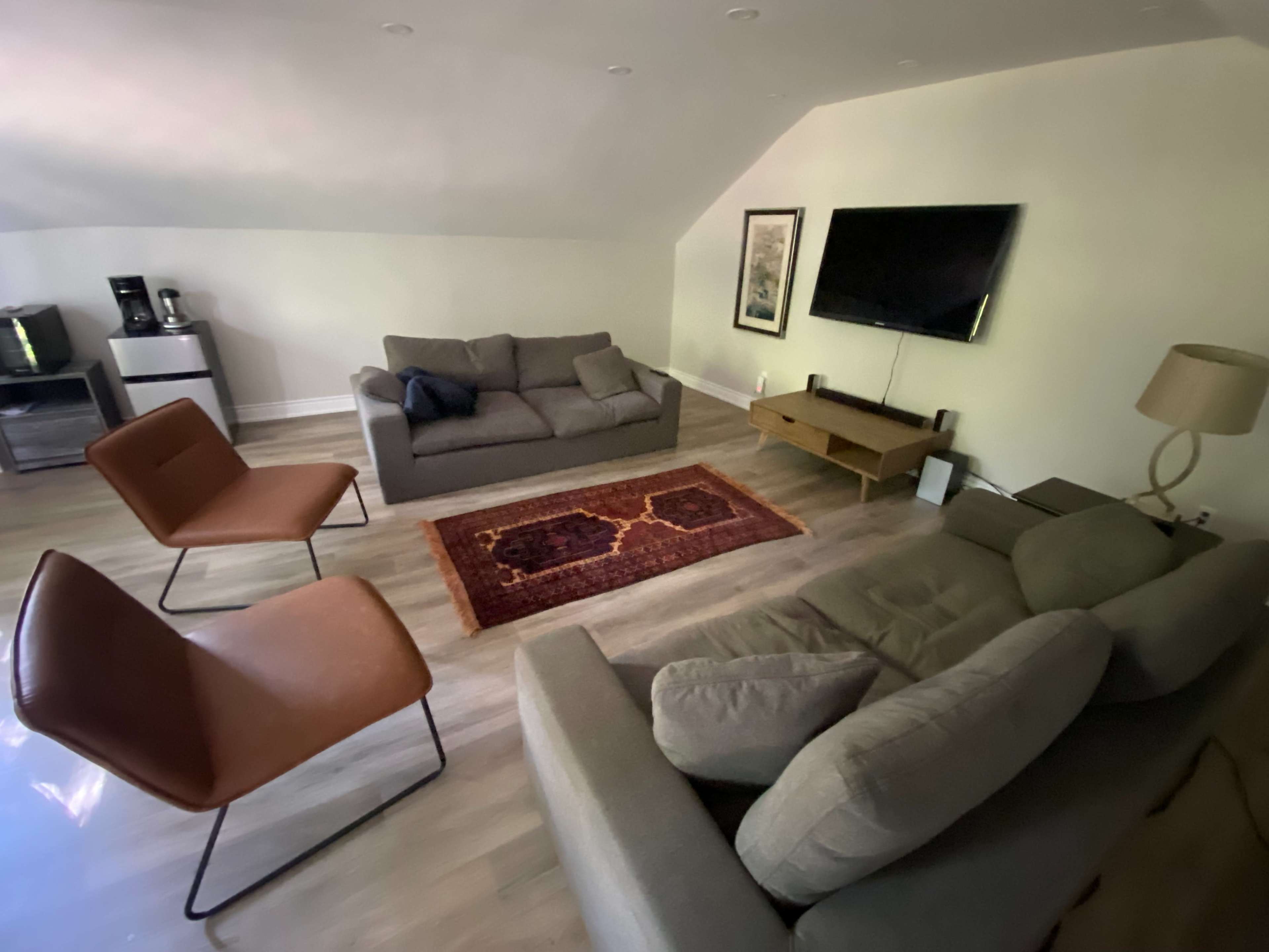 The image shows a cozy attic living space with a gray sofa, brown chairs, a television mounted on the wall, and a rug in the center.