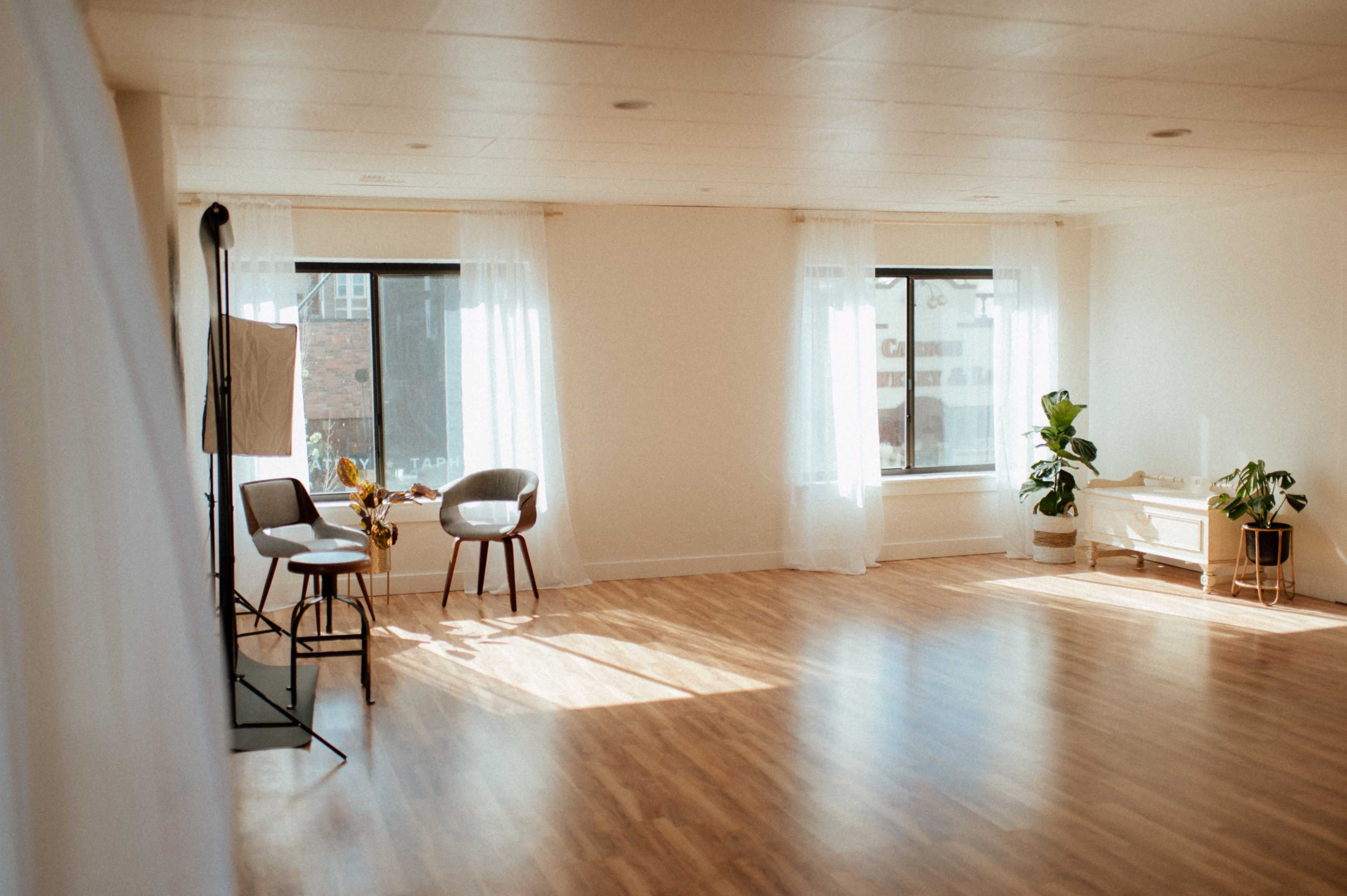 A bright and open studio space features two chairs, large windows with sheer curtains, and a potted plant.