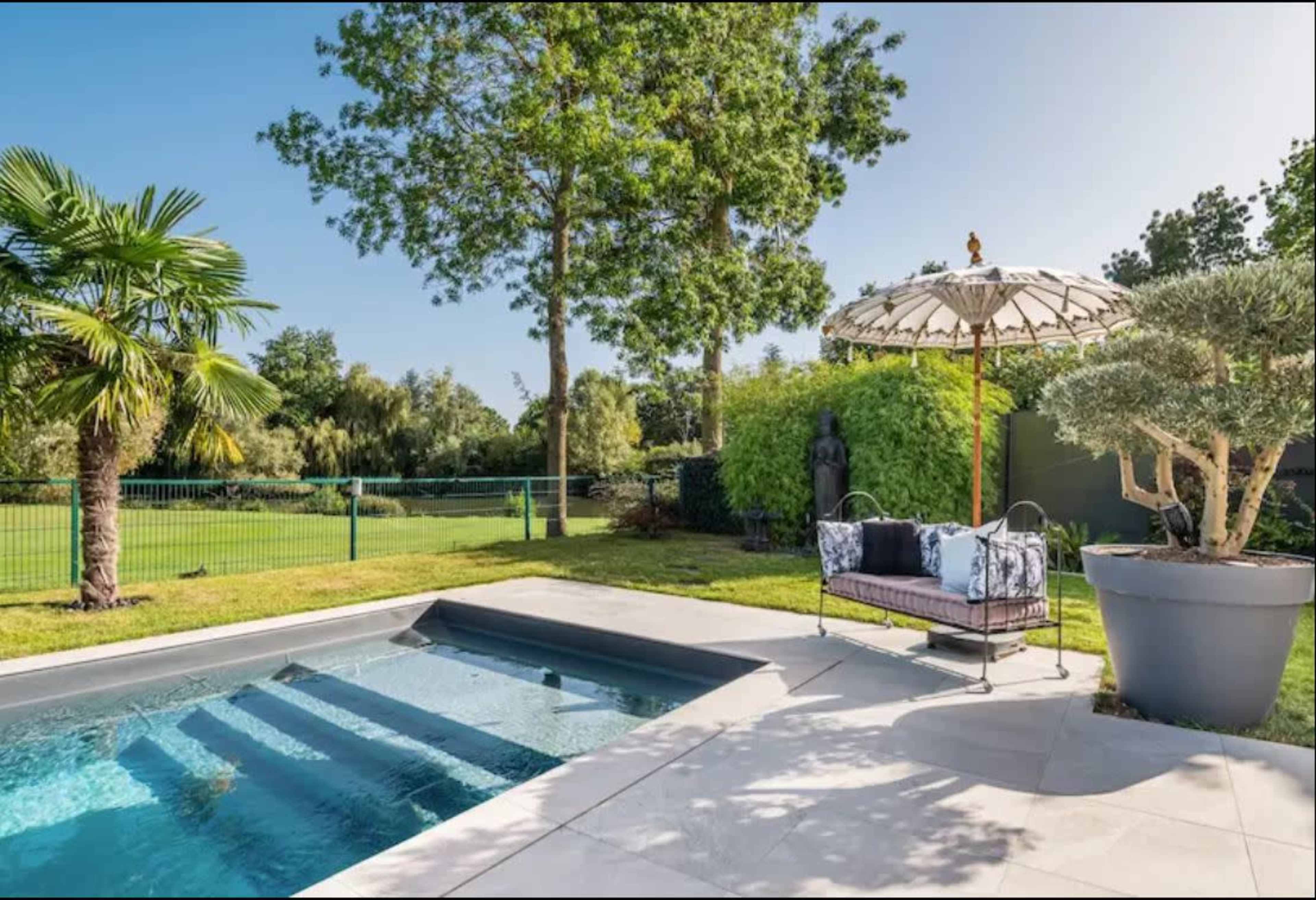 A modern pool area features a sleek pool beside a lounge seat and an umbrella, surrounded by greenery and trees.