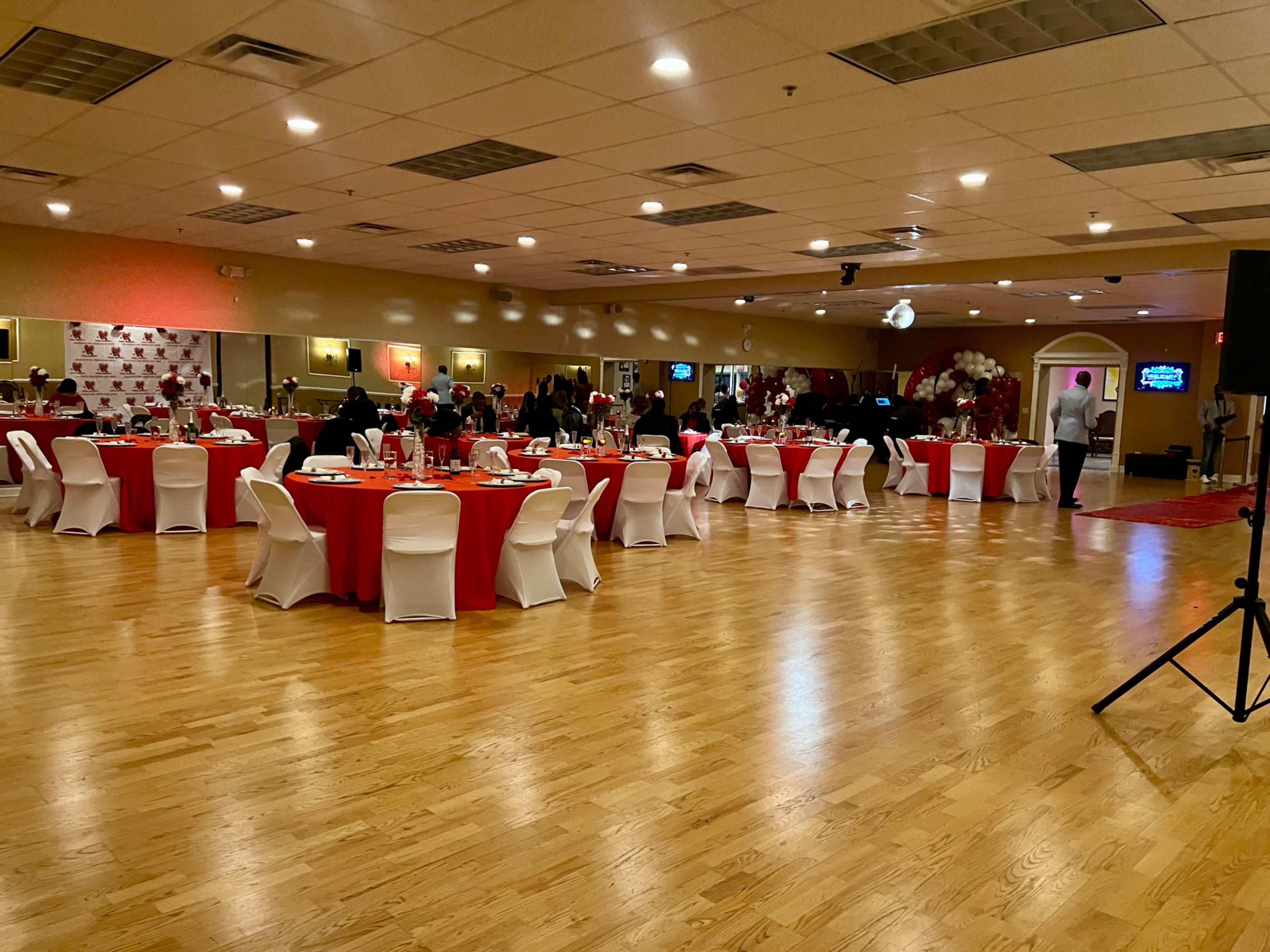 The image shows a spacious banquet hall filled with round tables covered in red tablecloths and surrounded by white chairs, where guests are engaged in various activities.