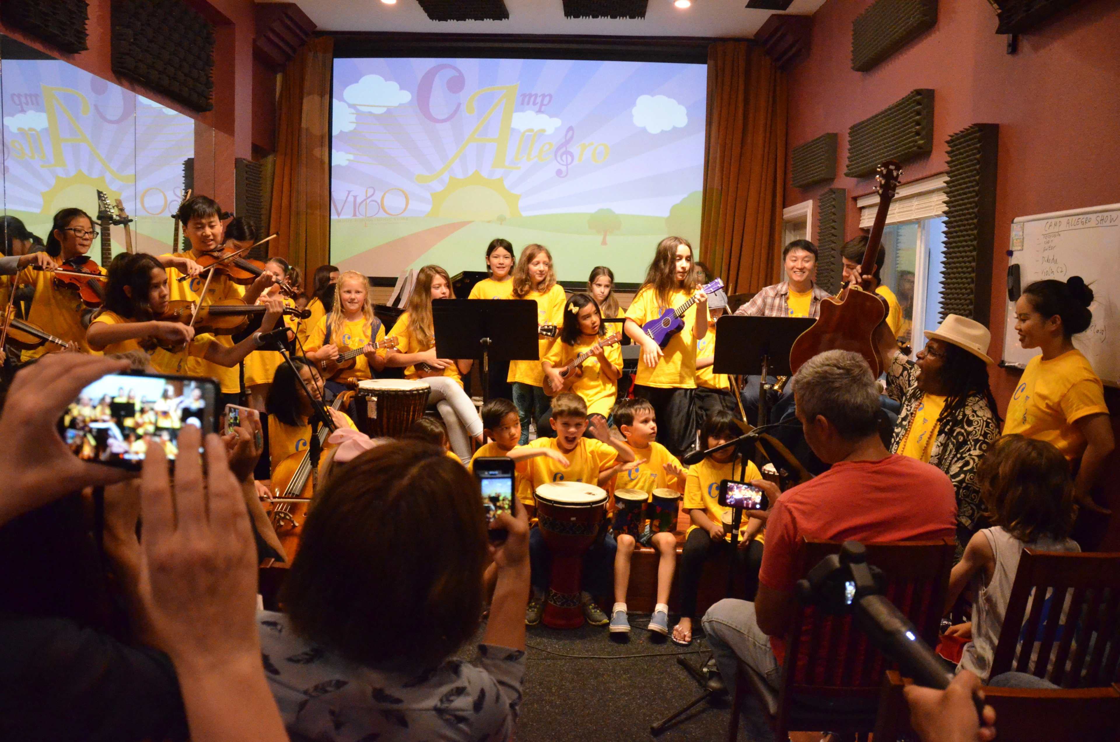 A large group of children in yellow shirts plays various musical instruments in a performance setting, while an audience records the event on their devices.