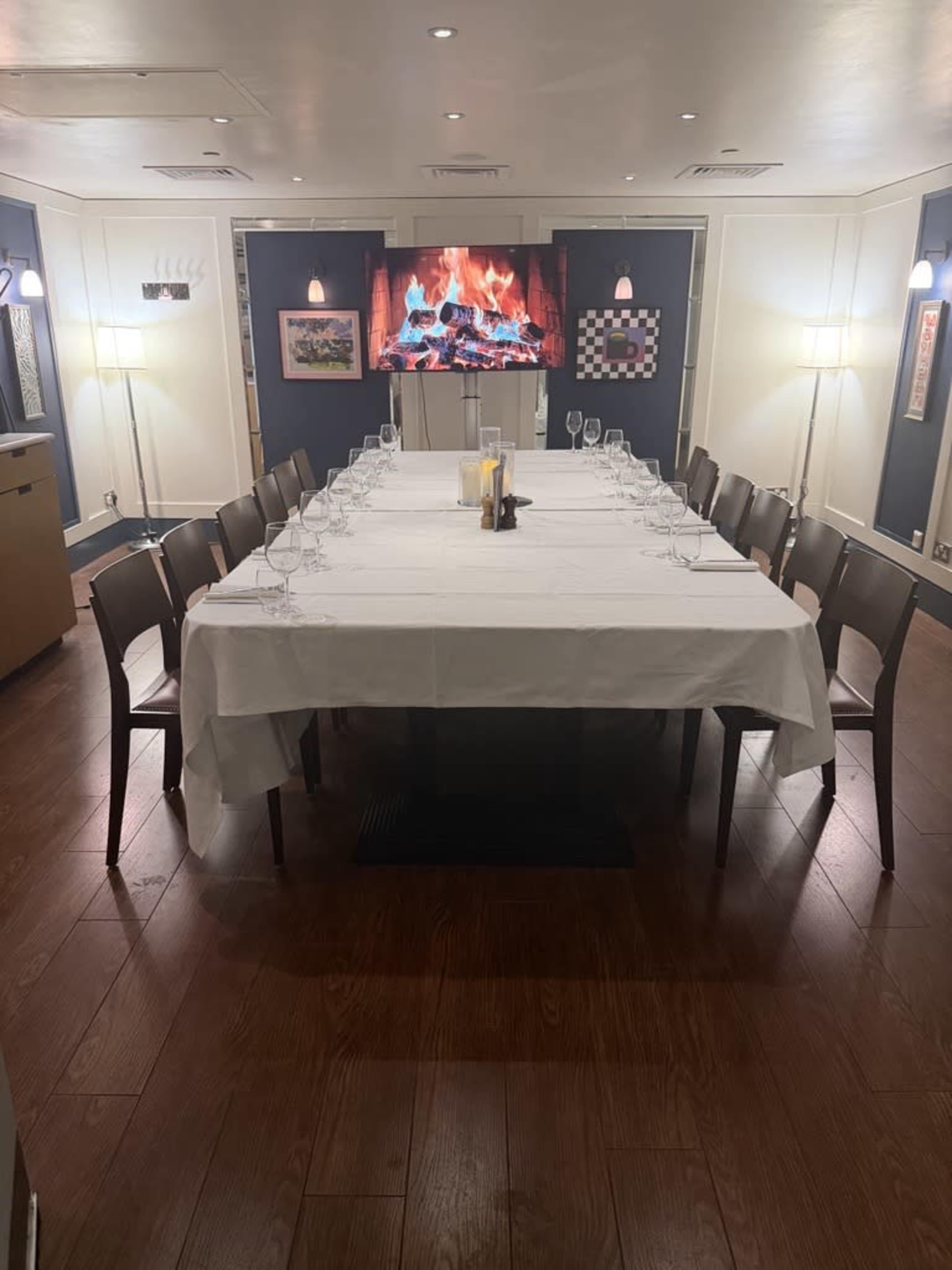 A long table set for dining is centrally placed in a well-lit room, with artwork on the walls and a fireplace video display in the background.
