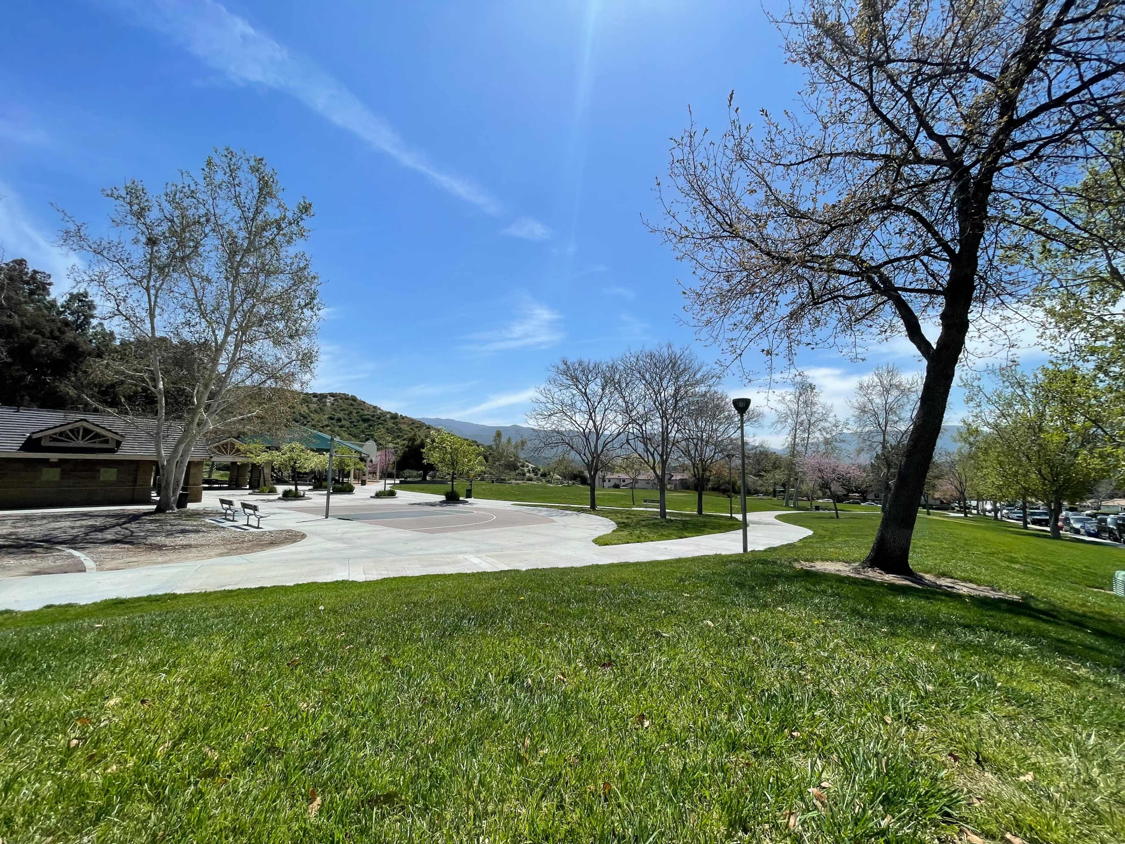 A paved pathway winds through a park with sparse trees and a backdrop of hills under a clear blue sky.
