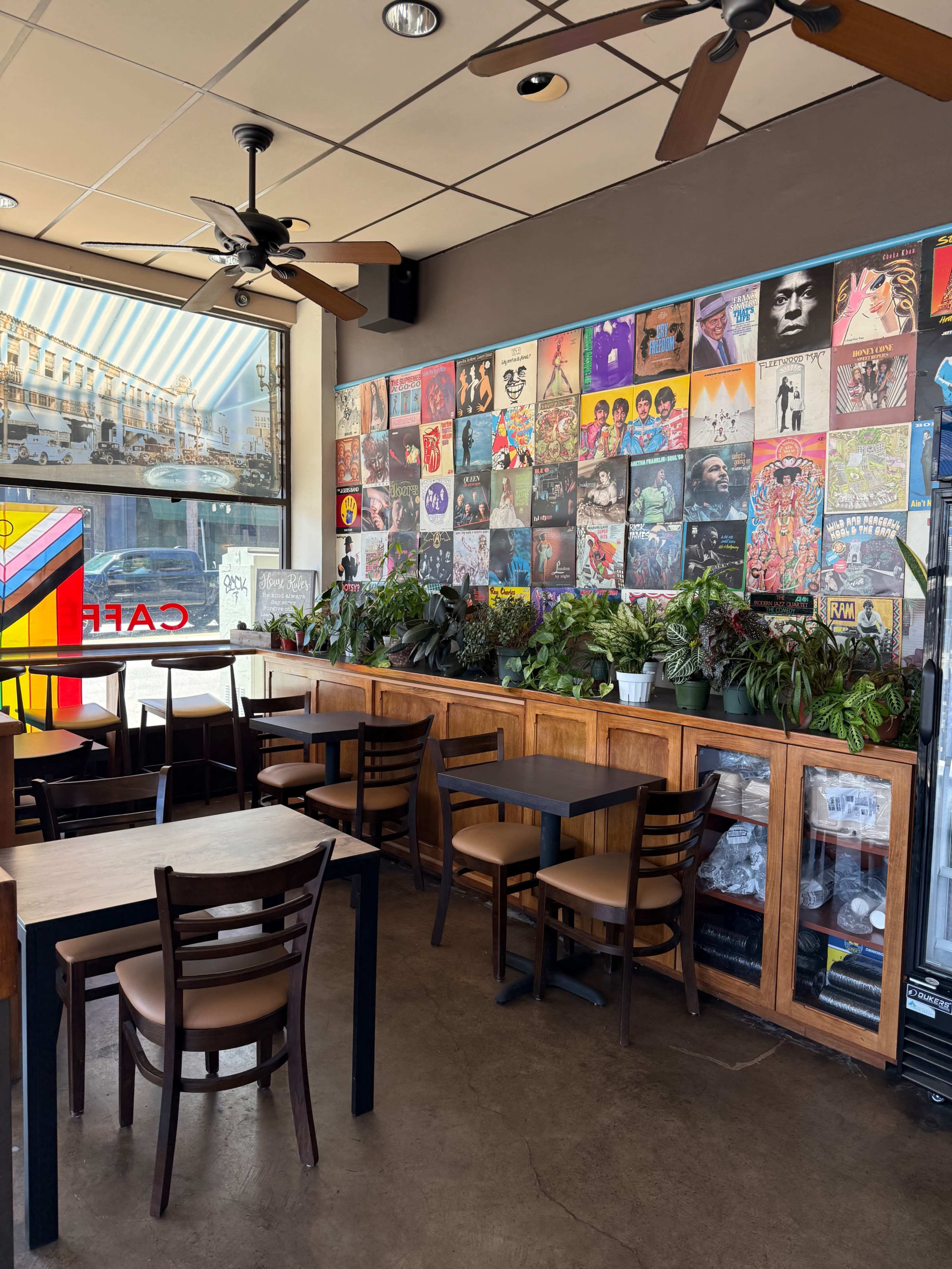 The interior of a café features wooden tables and chairs arranged near a wall decorated with various colorful album covers and houseplants.