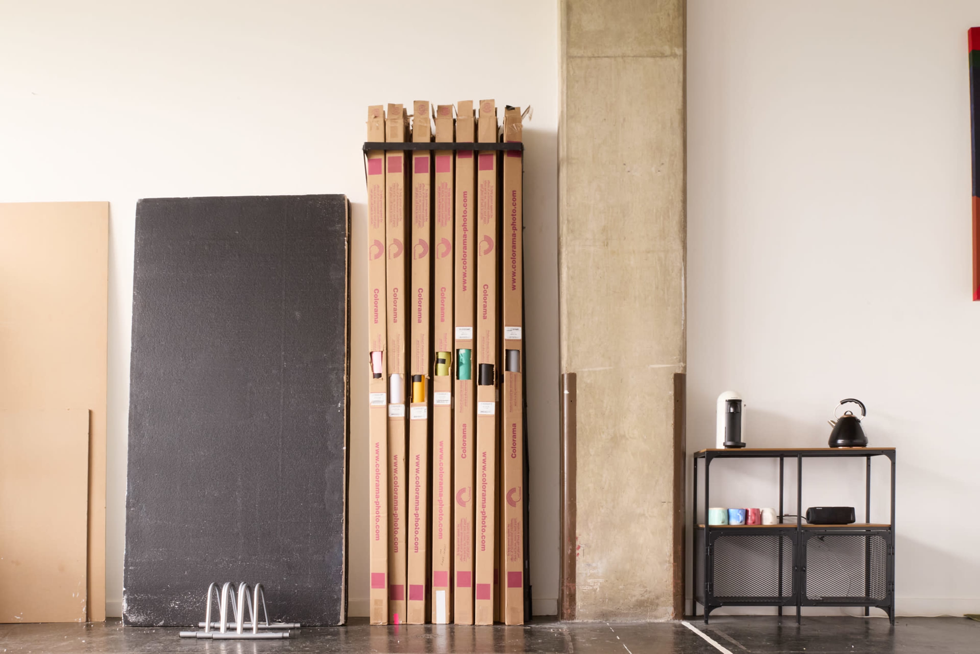 Several large, flattened cardboard boxes stacked against a wall alongside a black storage cart and large sheets of material on the floor.