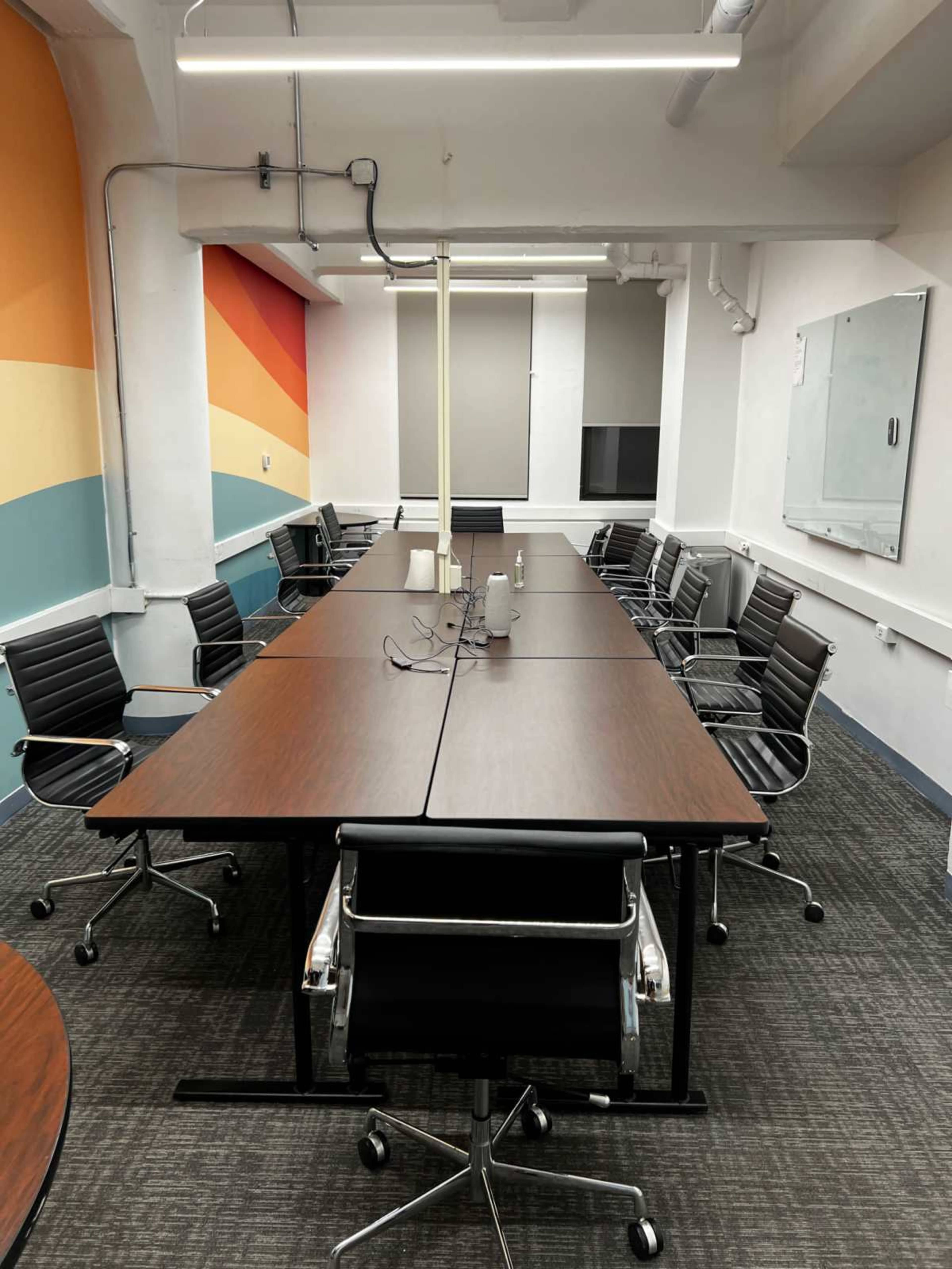The image shows a large conference room with several black ergonomic chairs arranged around rectangular wooden tables and a colorful wall mural in the background.