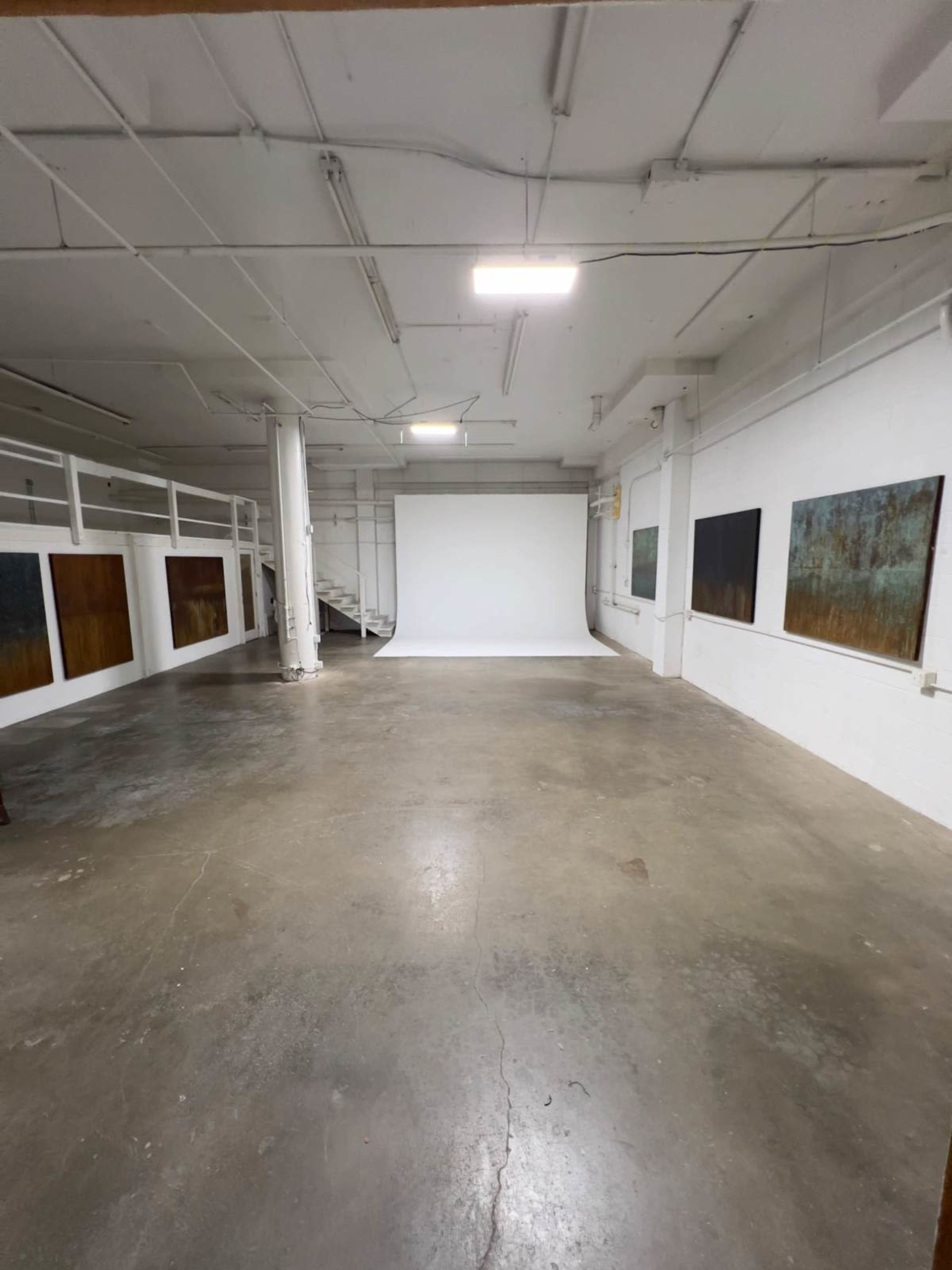 The image shows an empty industrial-style room with a concrete floor, white walls, and a blank backdrop at one end.