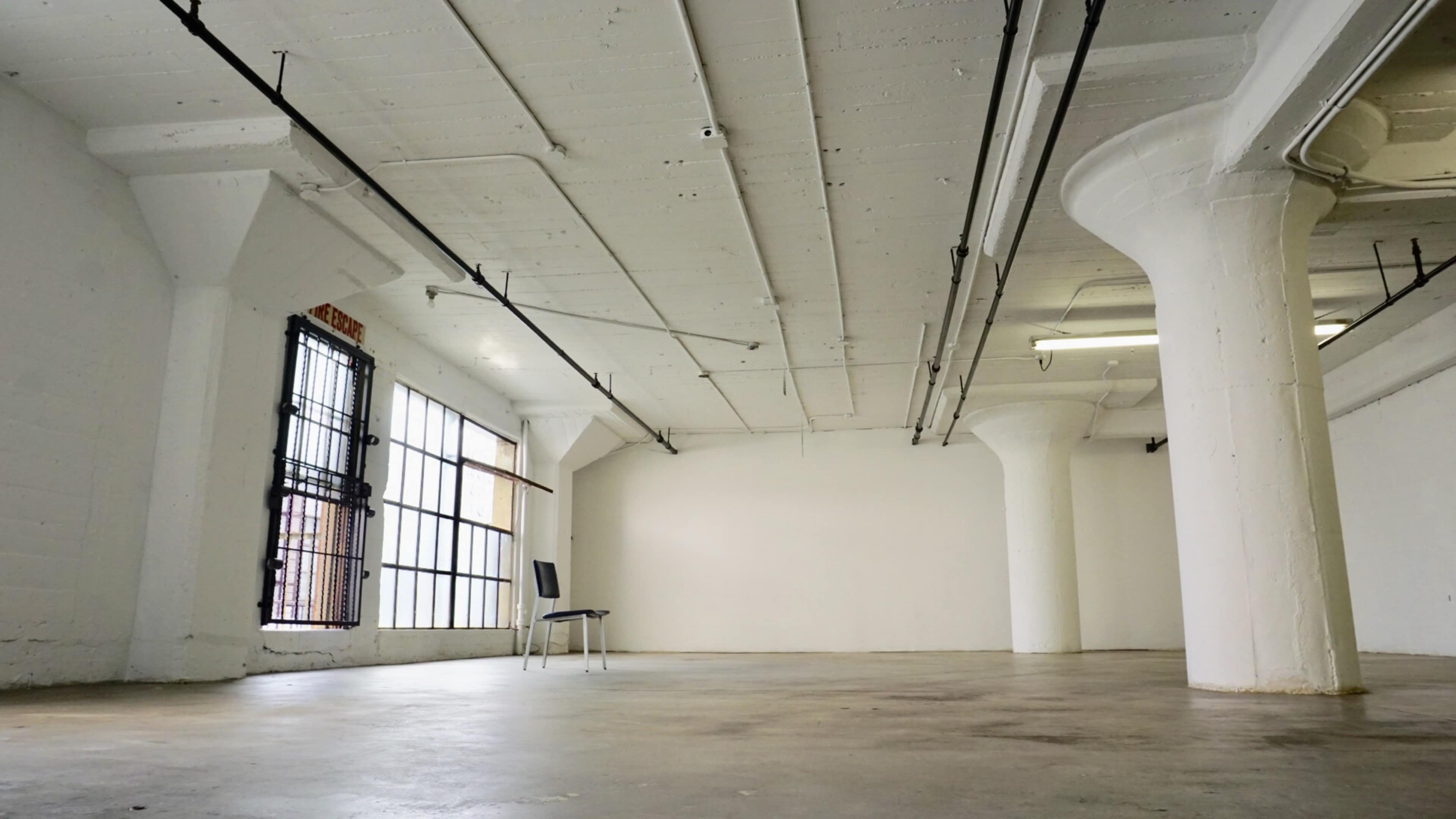 The image shows an empty, spacious room with a single chair positioned against a white wall and large windows letting in natural light.