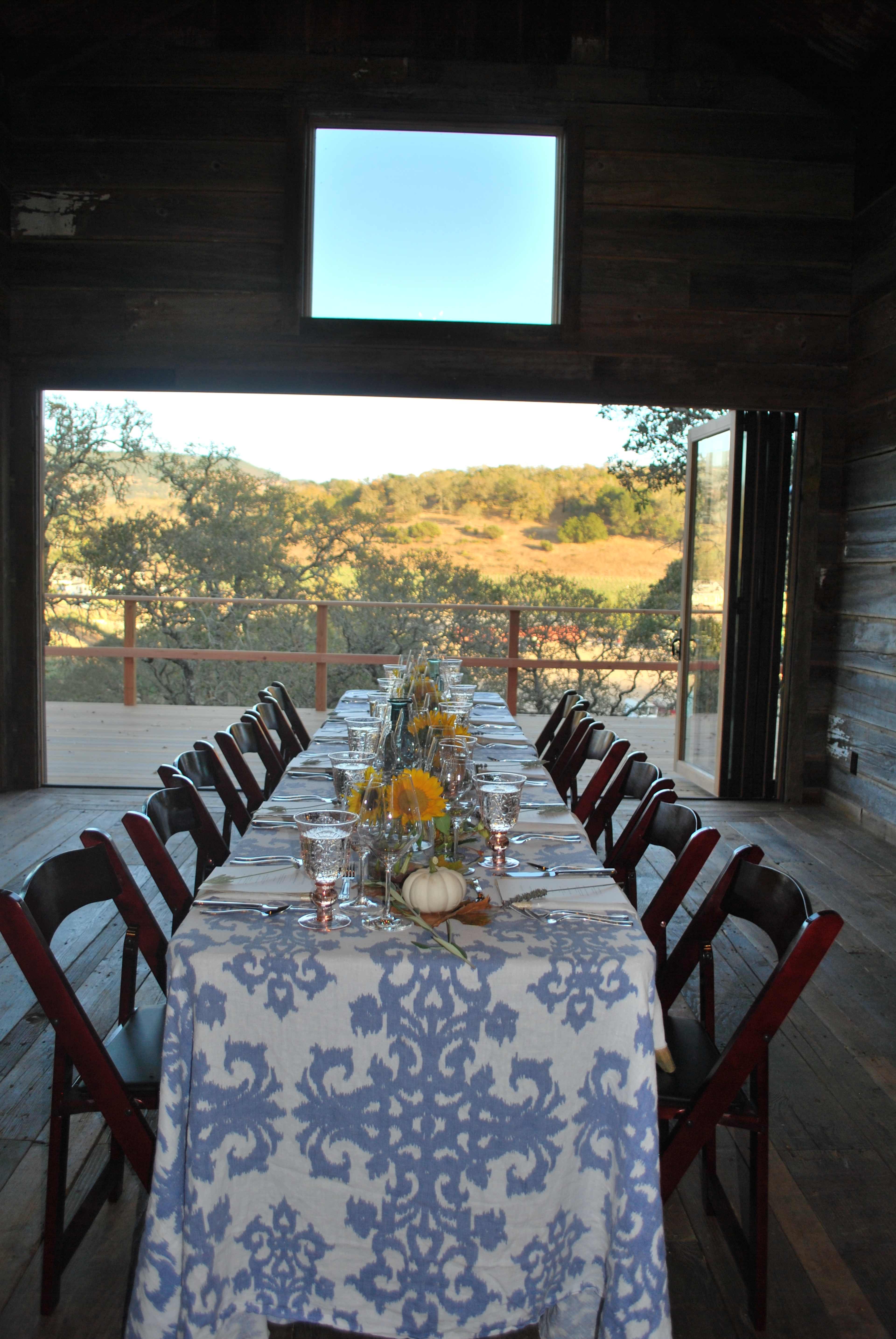 A long dining table is set with a floral centerpiece and is positioned in front of an open doorway that leads to a view of a hillside.