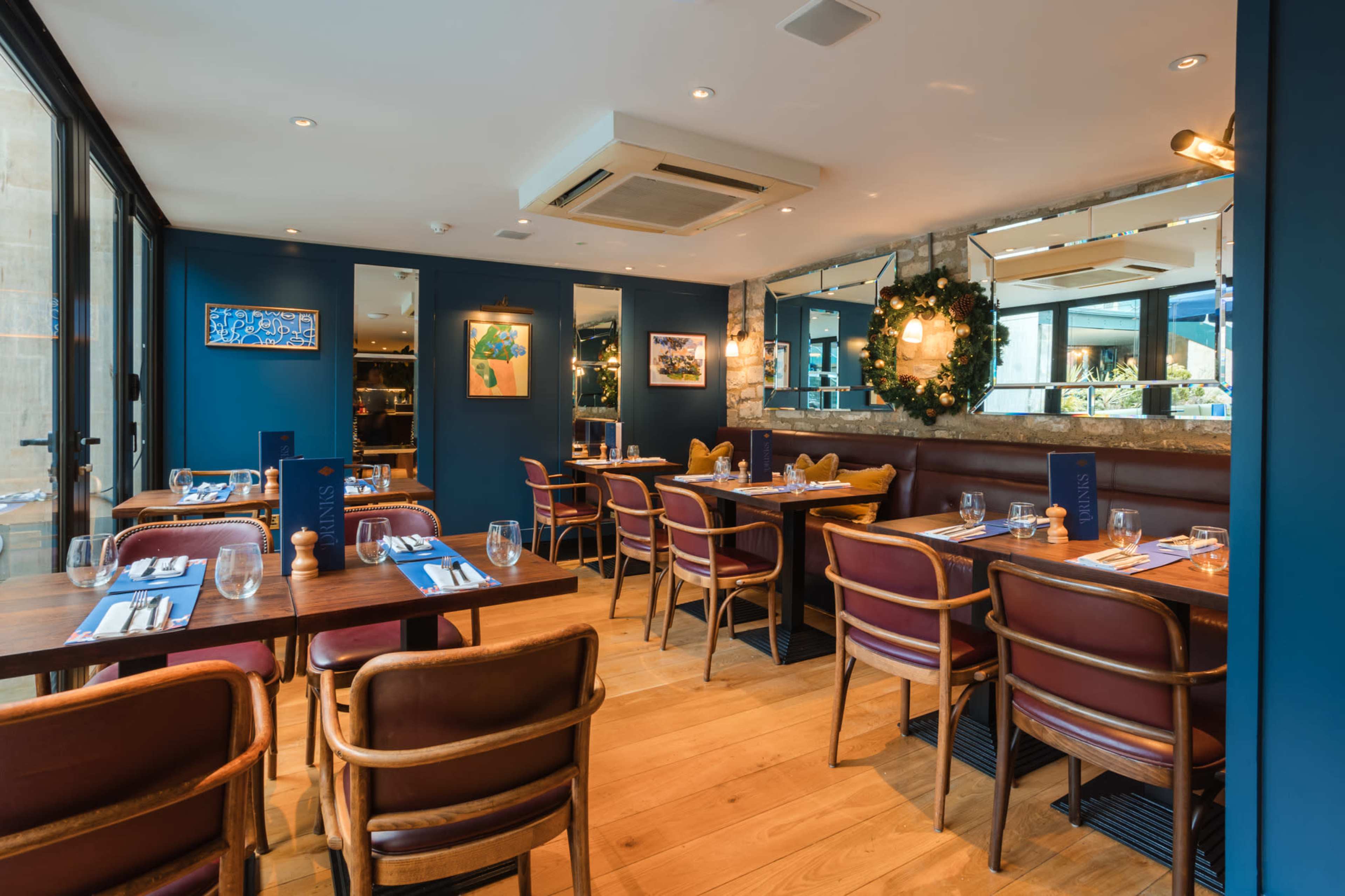 The image shows a modern restaurant interior featuring wooden tables, upholstered chairs, and blue walls adorned with artwork and a mirror.