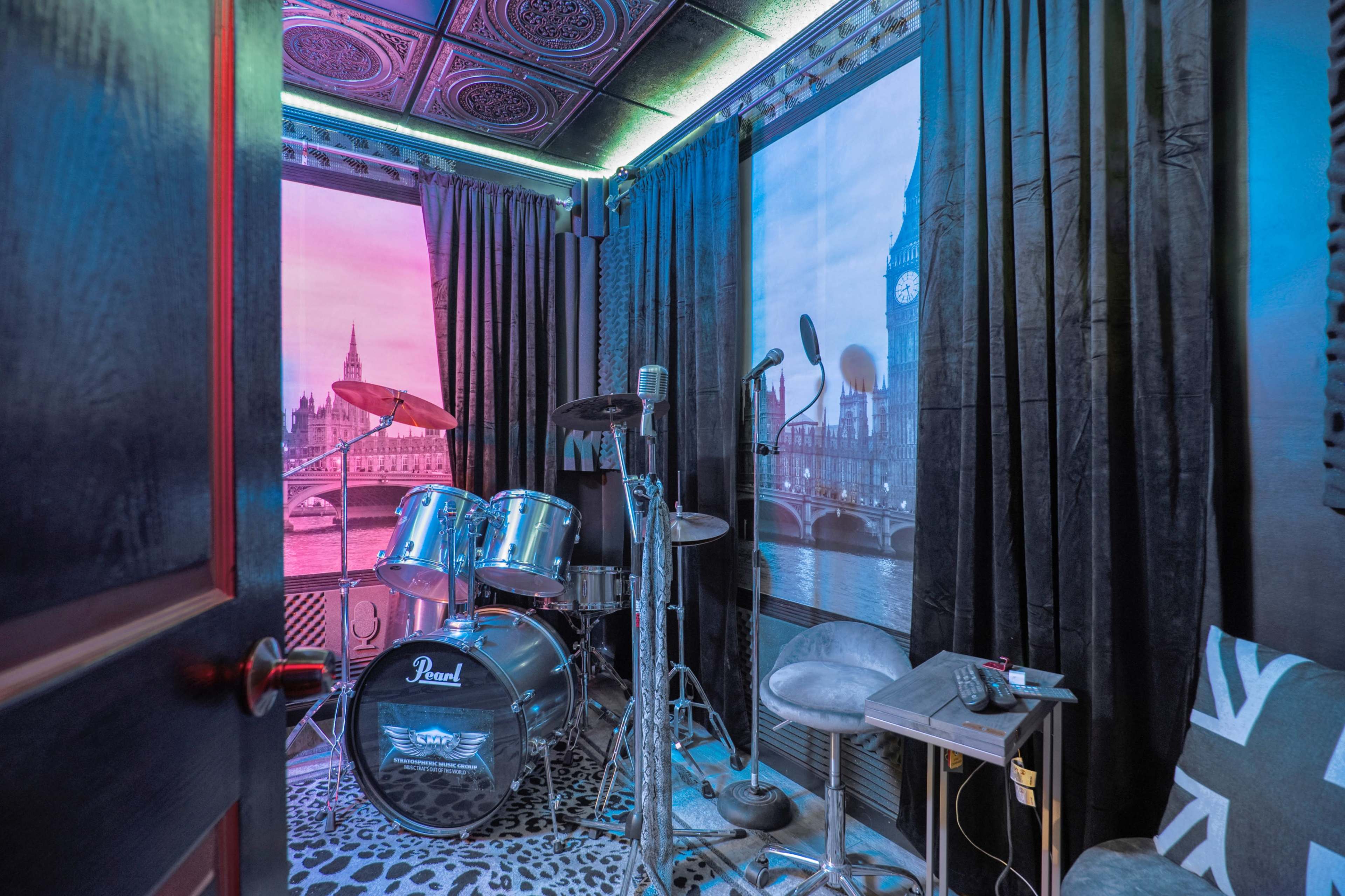 A small music rehearsal room featuring a drum set, microphones, and photos of London landmarks on the walls.