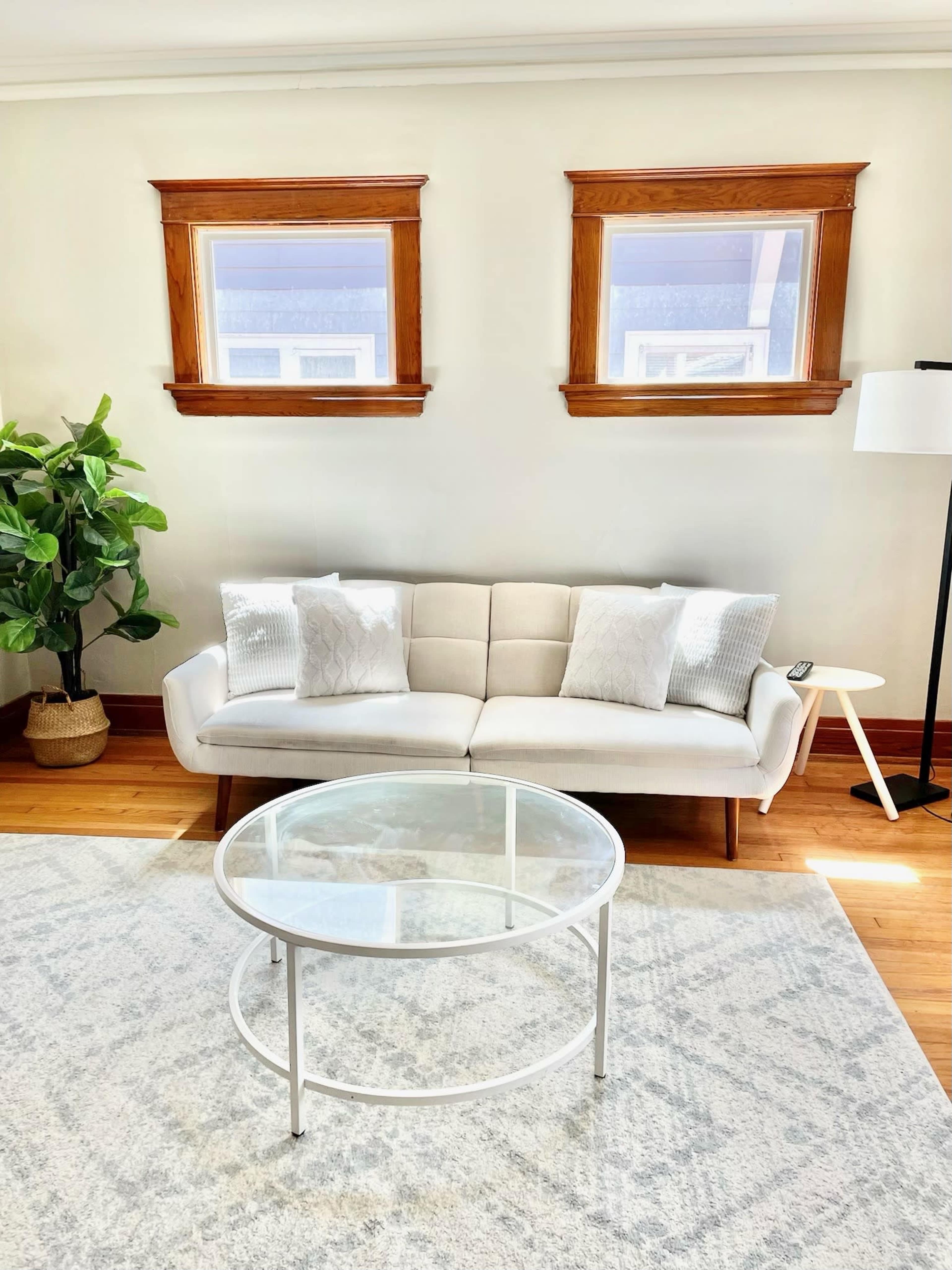 A beige sofa with decorative pillows is positioned in a bright room featuring two windows, a potted plant, and a round glass coffee table on a light area rug.