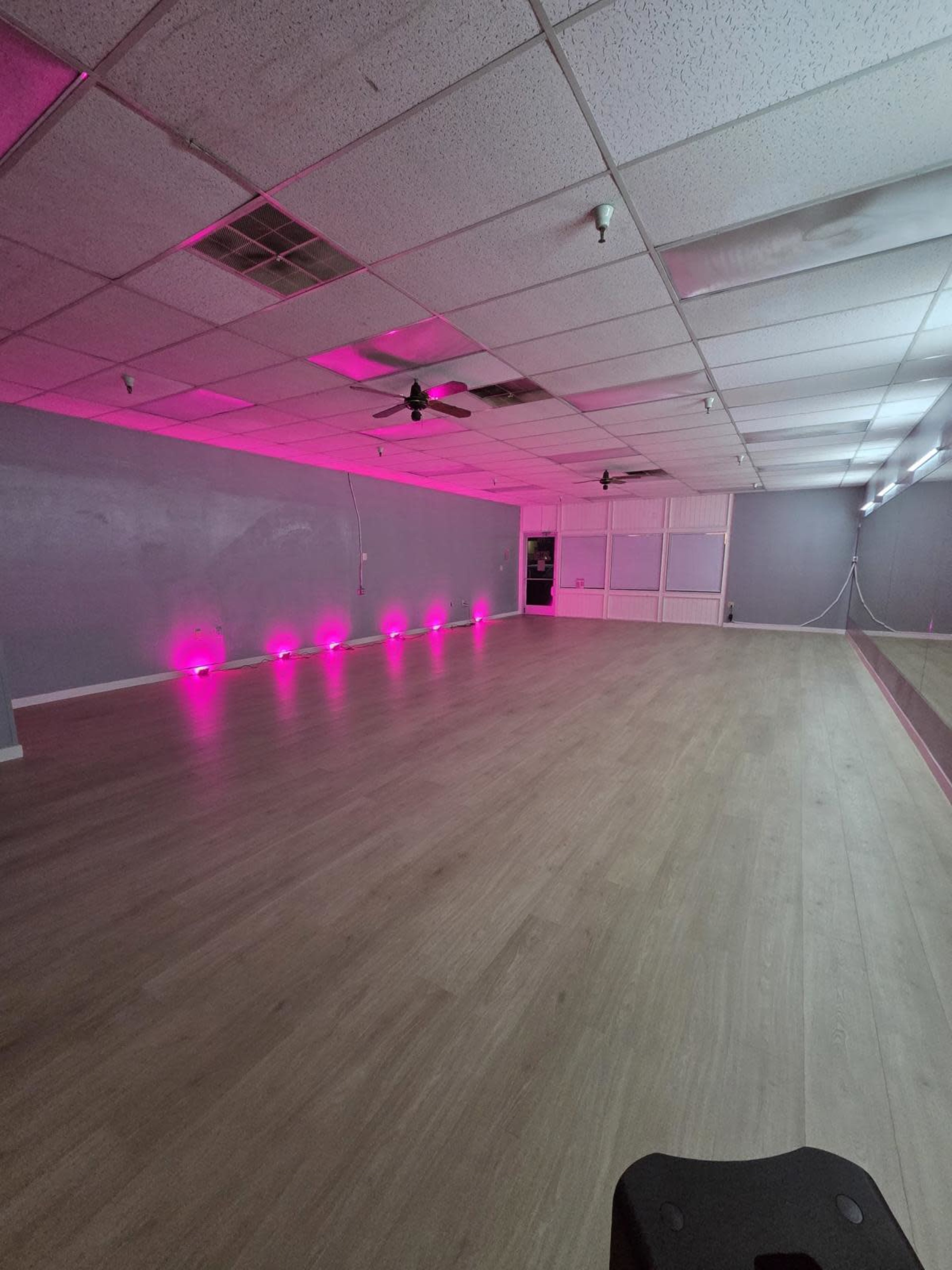 The image shows an empty room with wooden flooring, bright pink lighting along the walls, and a large mirror on one side.