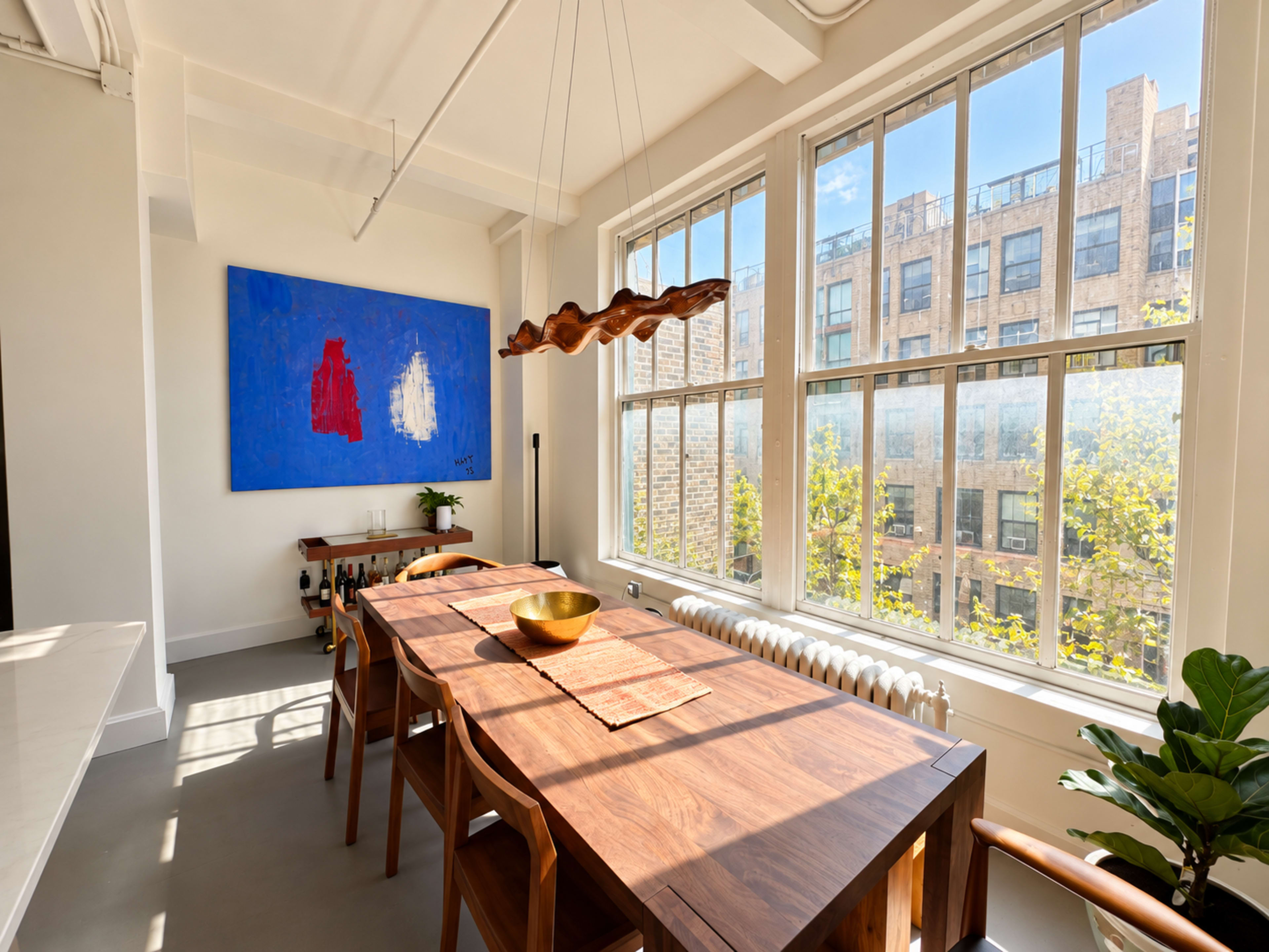 A dining area features a long wooden table, large windows allowing natural light, and modern artwork on the wall.