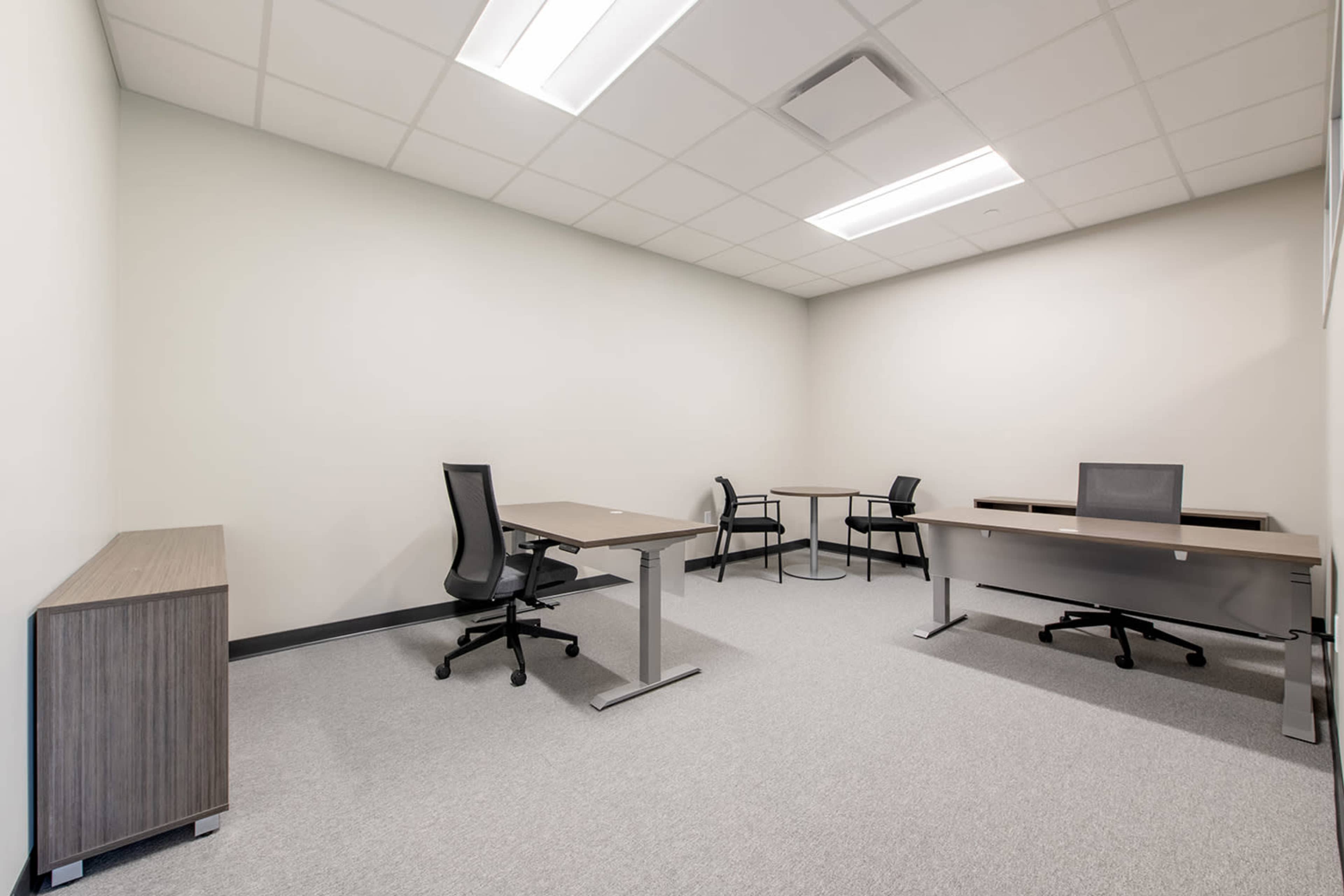 The image shows a simple office space featuring two desks and four chairs arranged in a minimalistic layout, with plain walls and carpeted flooring.