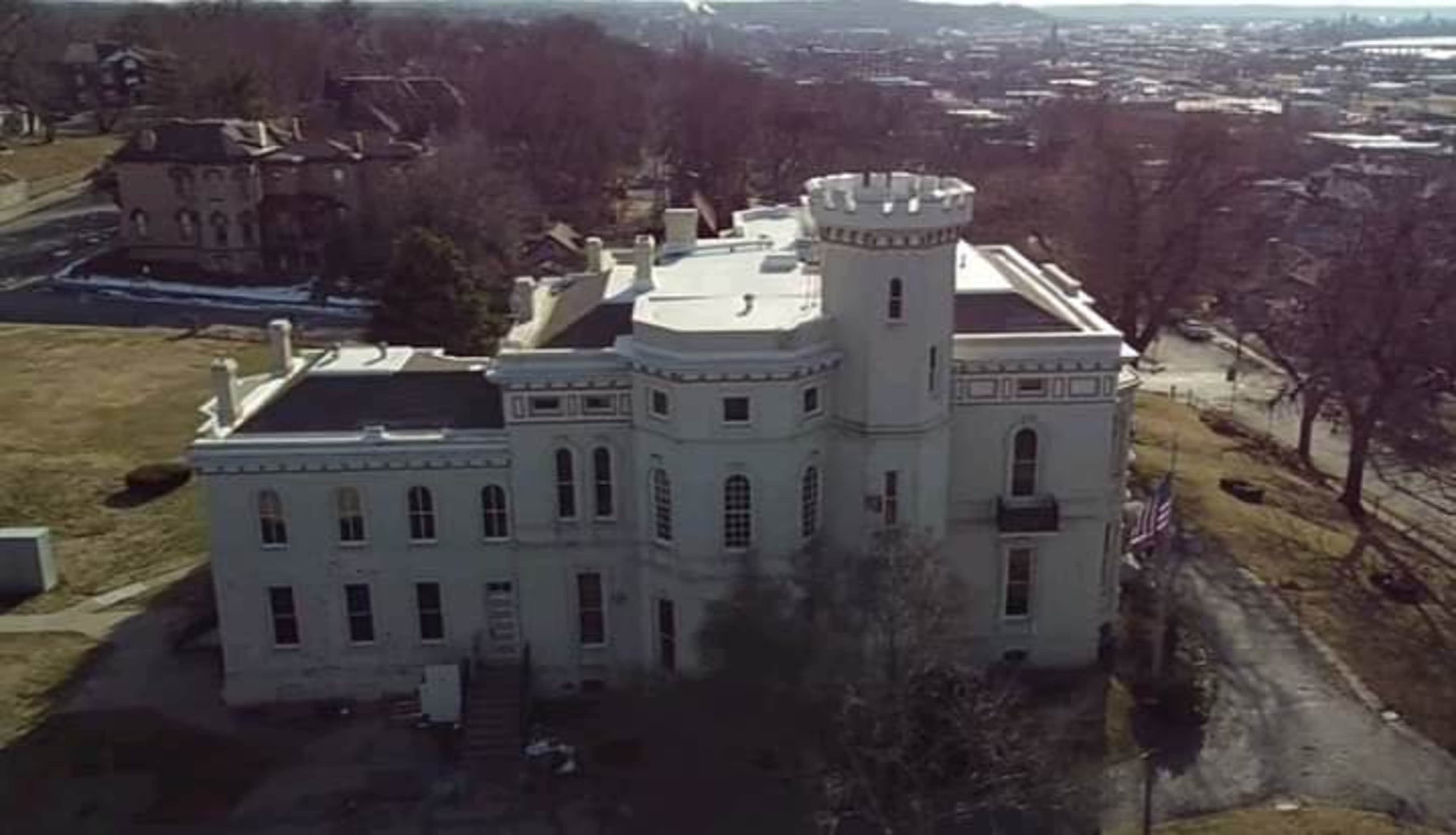 The image shows a large white historical building with a turret and an adjacent grassy area, surrounded by trees and a view of a city in the background.