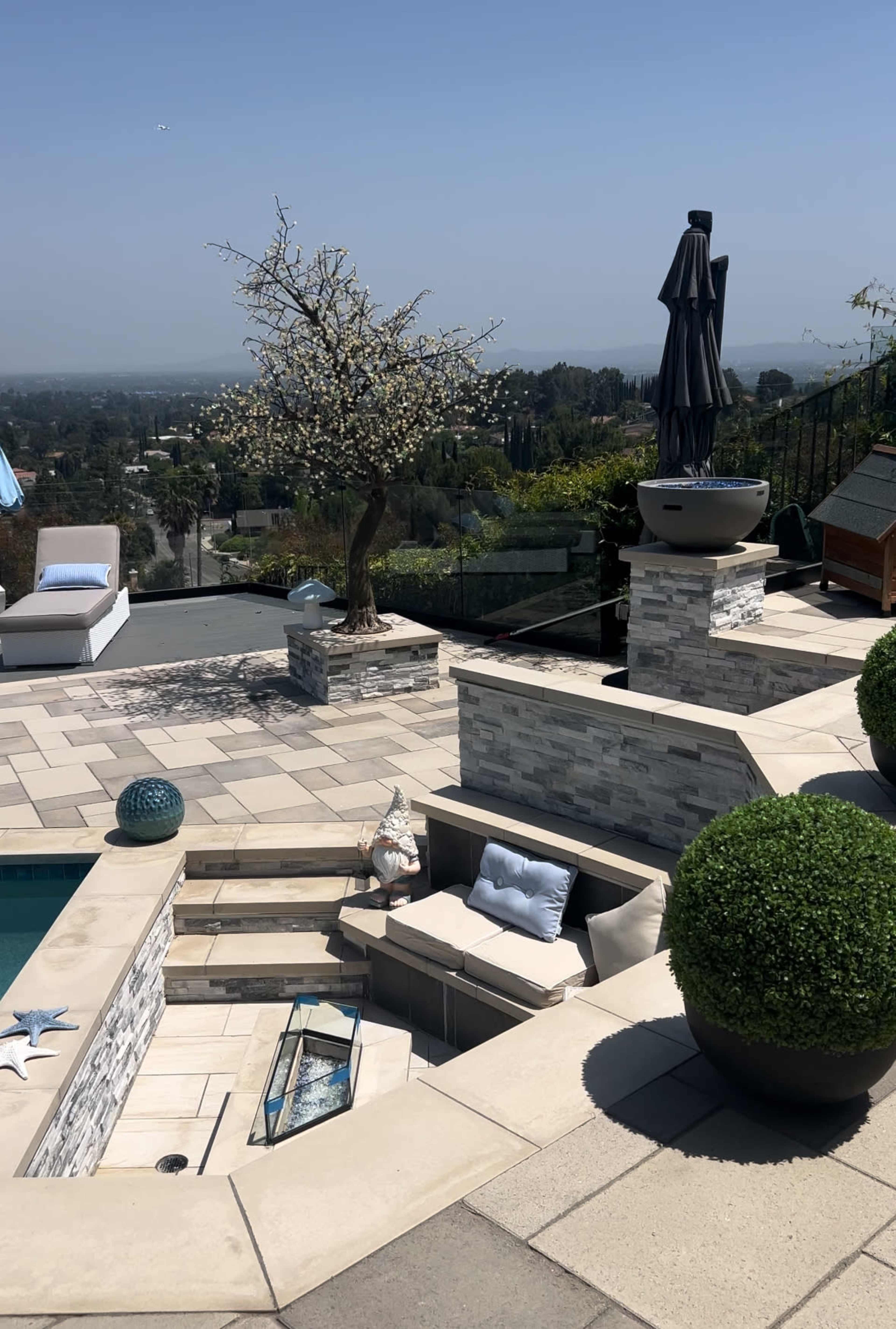 A landscaped outdoor area with a pool, stone seating, a statue, and trimmed bushes overlooking a valley.