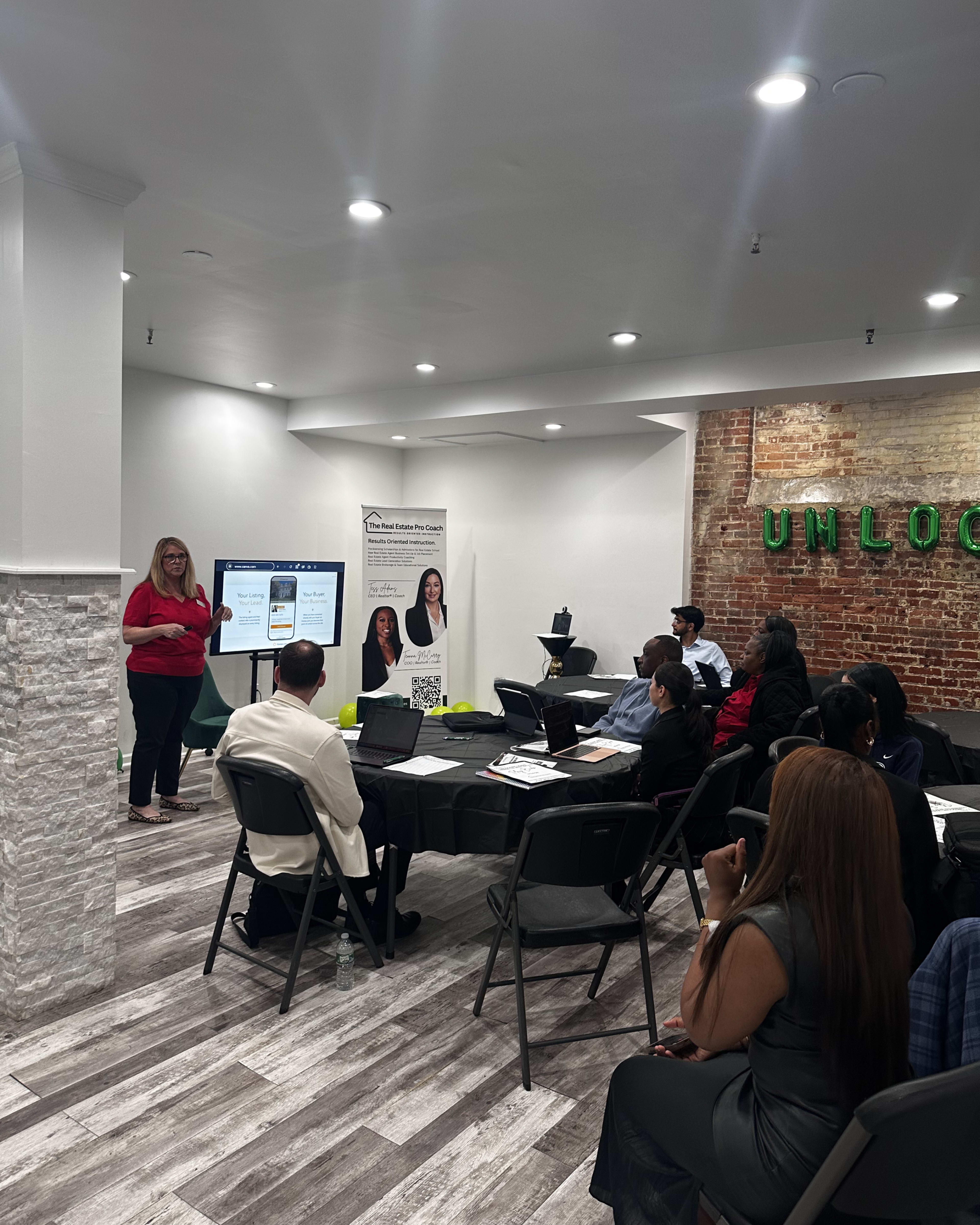 A presenter stands in front of a group seated at tables, showcasing a digital presentation in a well-lit room with exposed brick walls.