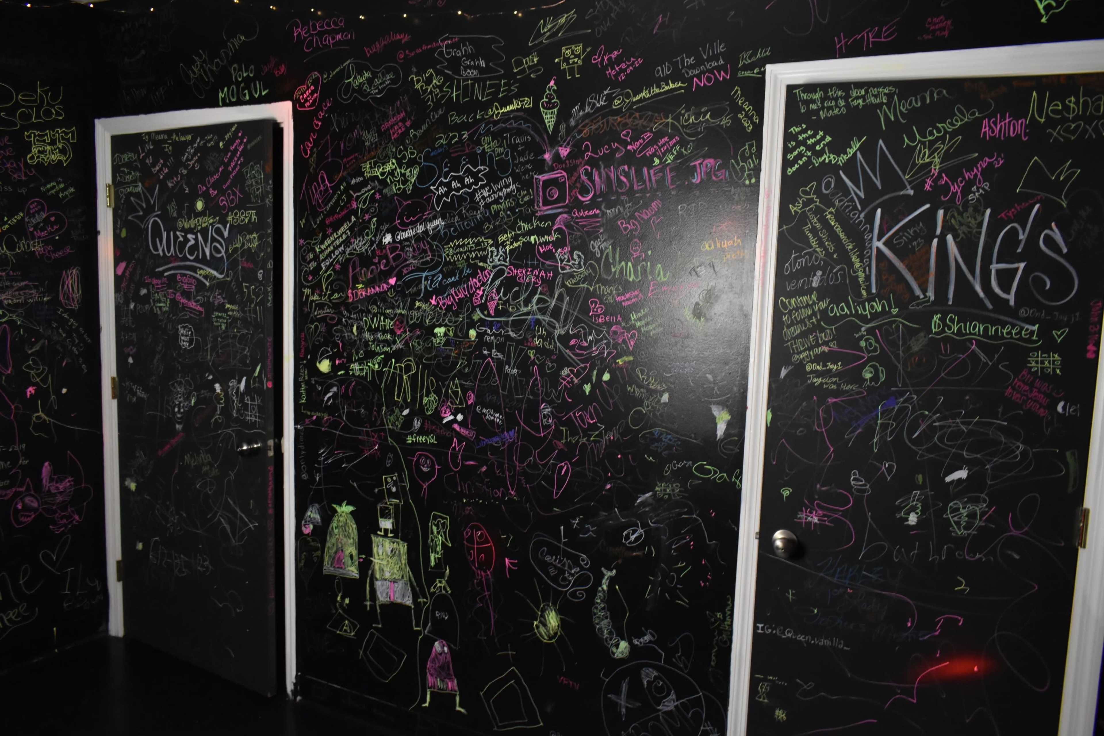 A dark room with two doors labeled "QUEENS" and "KINGS," and walls covered in colorful graffiti and drawings.