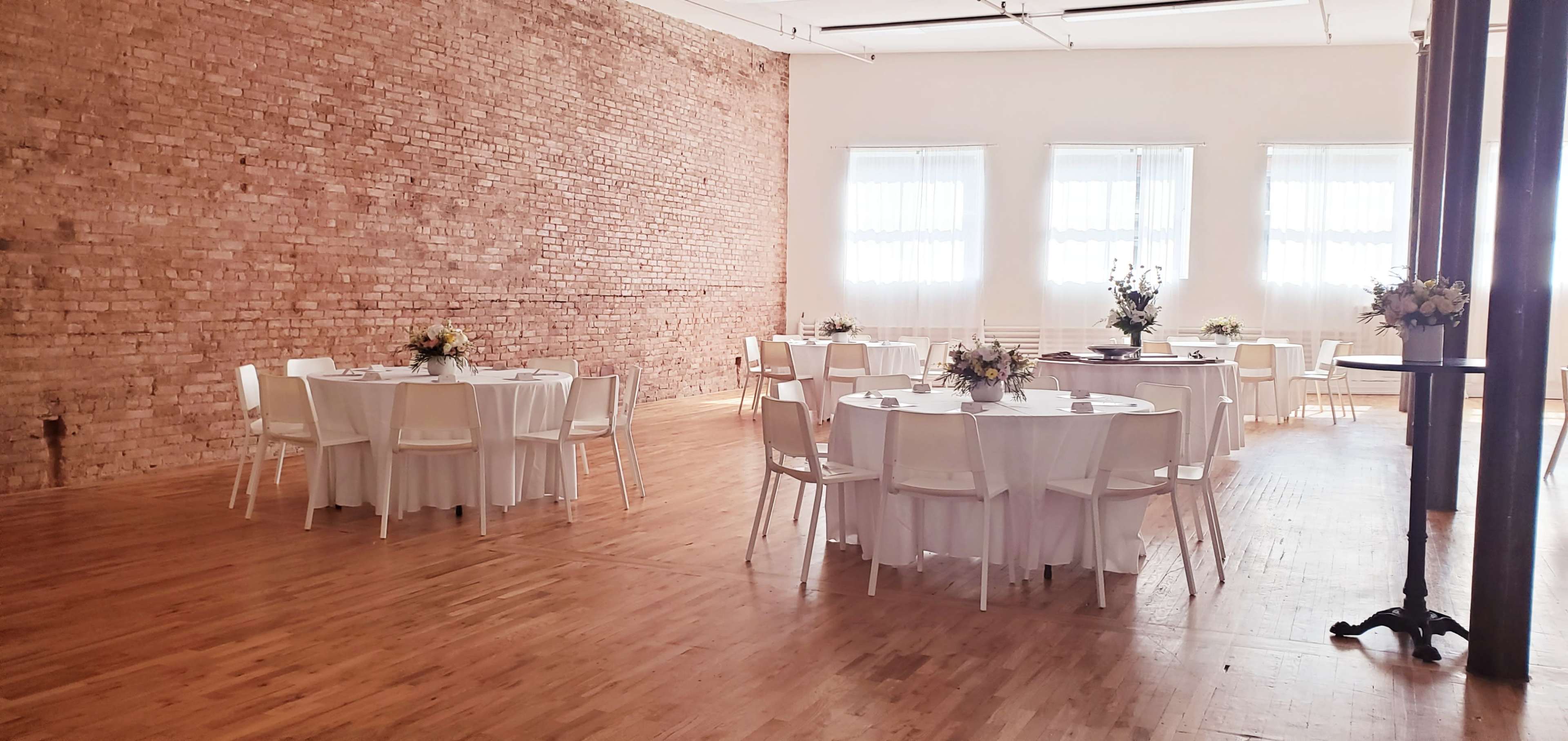 The scene shows a spacious event room with several round tables covered in white tablecloths, surrounded by chairs, set against a brick wall and bright windows.