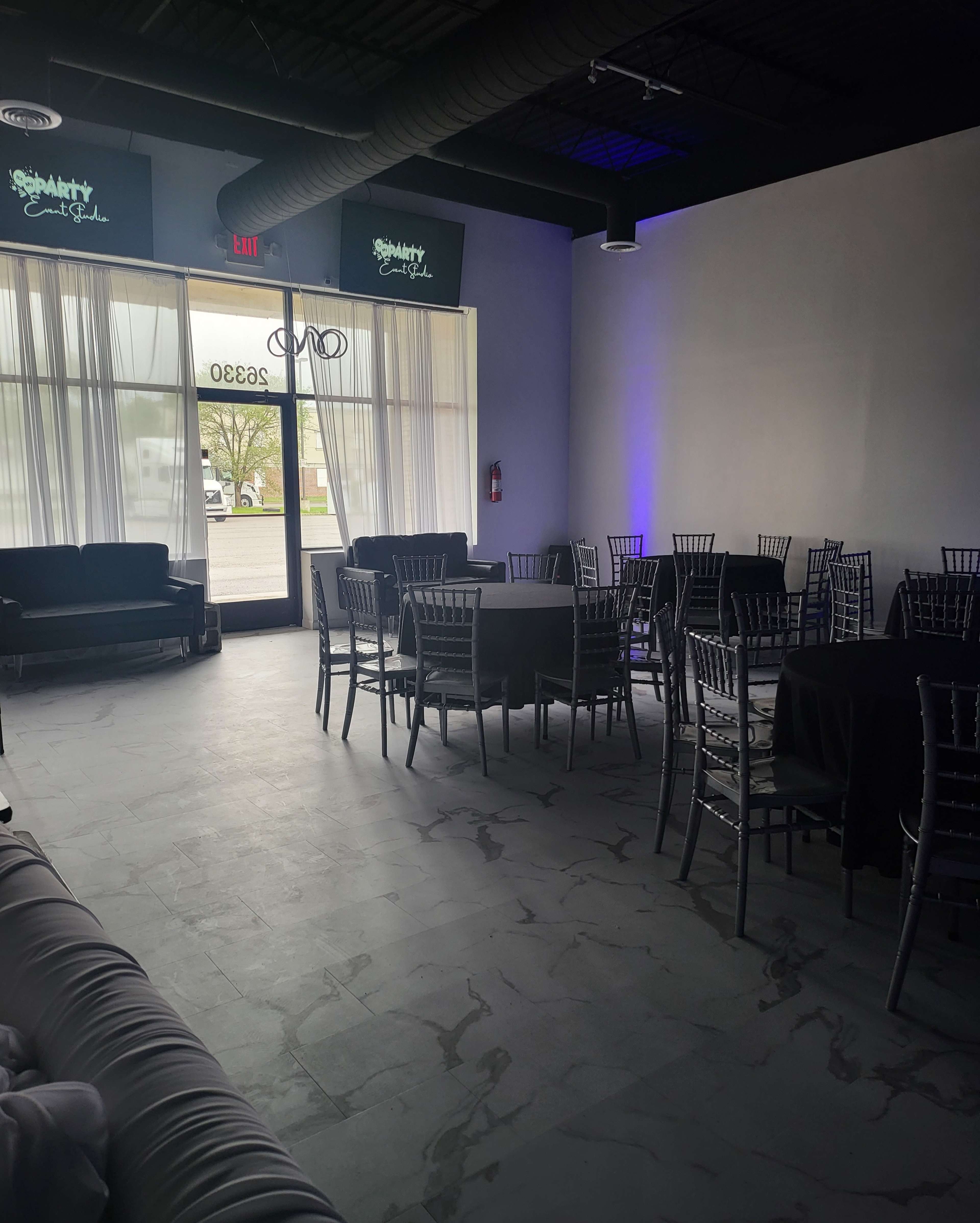 A spacious event venue with black tables and silver chiavari chairs, illuminated by natural light from large windows.