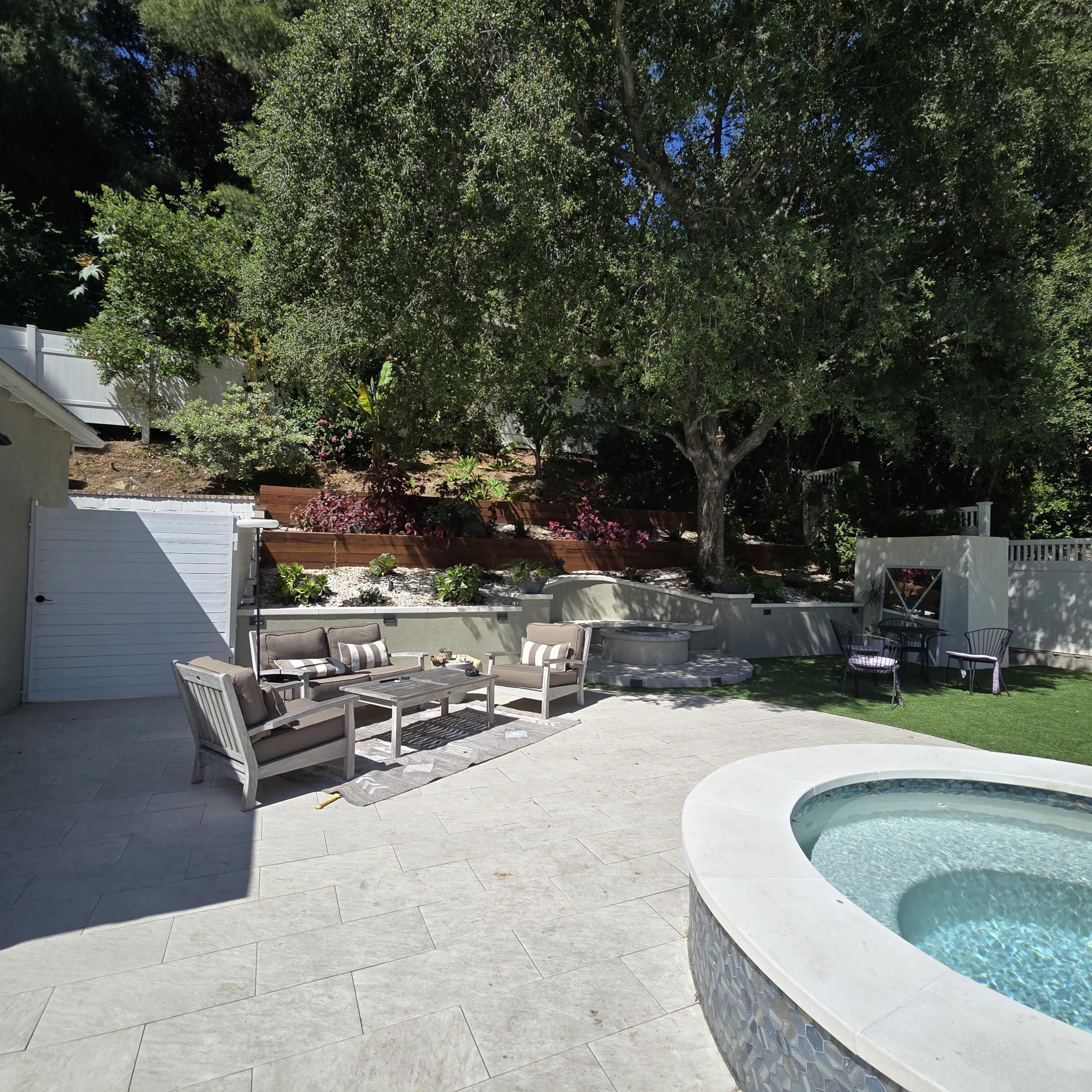 The image shows a landscaped backyard featuring seating on a patio, a hot tub, and a large tree surrounded by greenery.