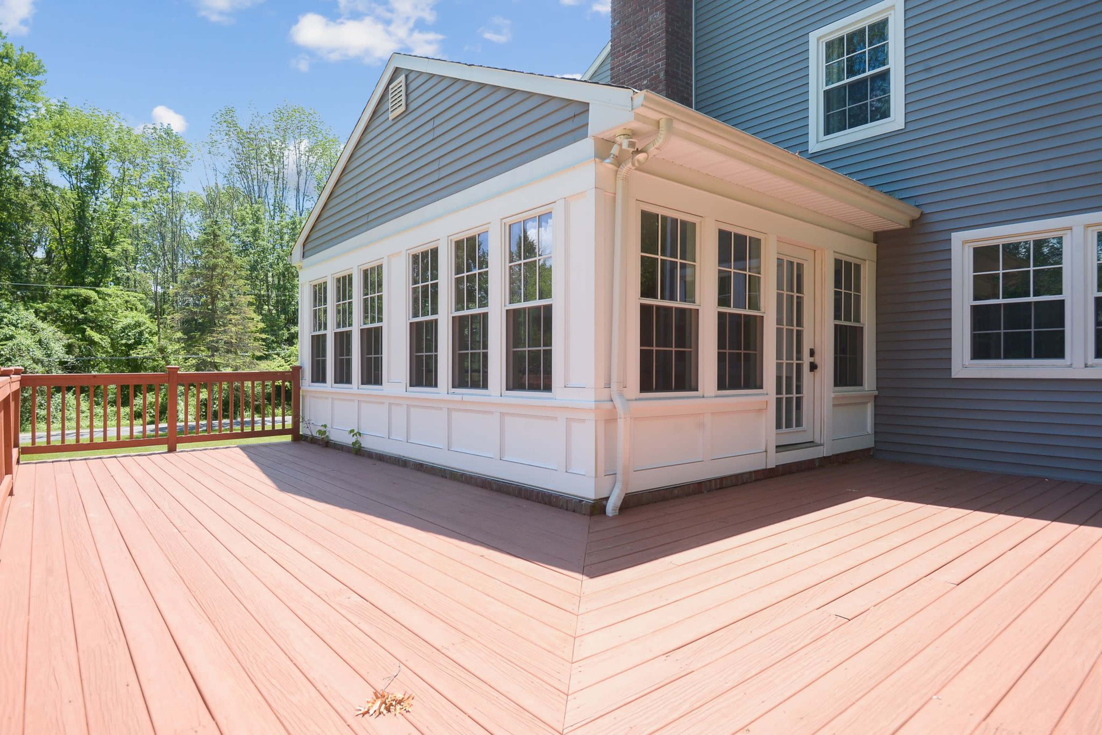 The image shows a wooden deck leading up to a gray and white sunroom with multiple windows.
