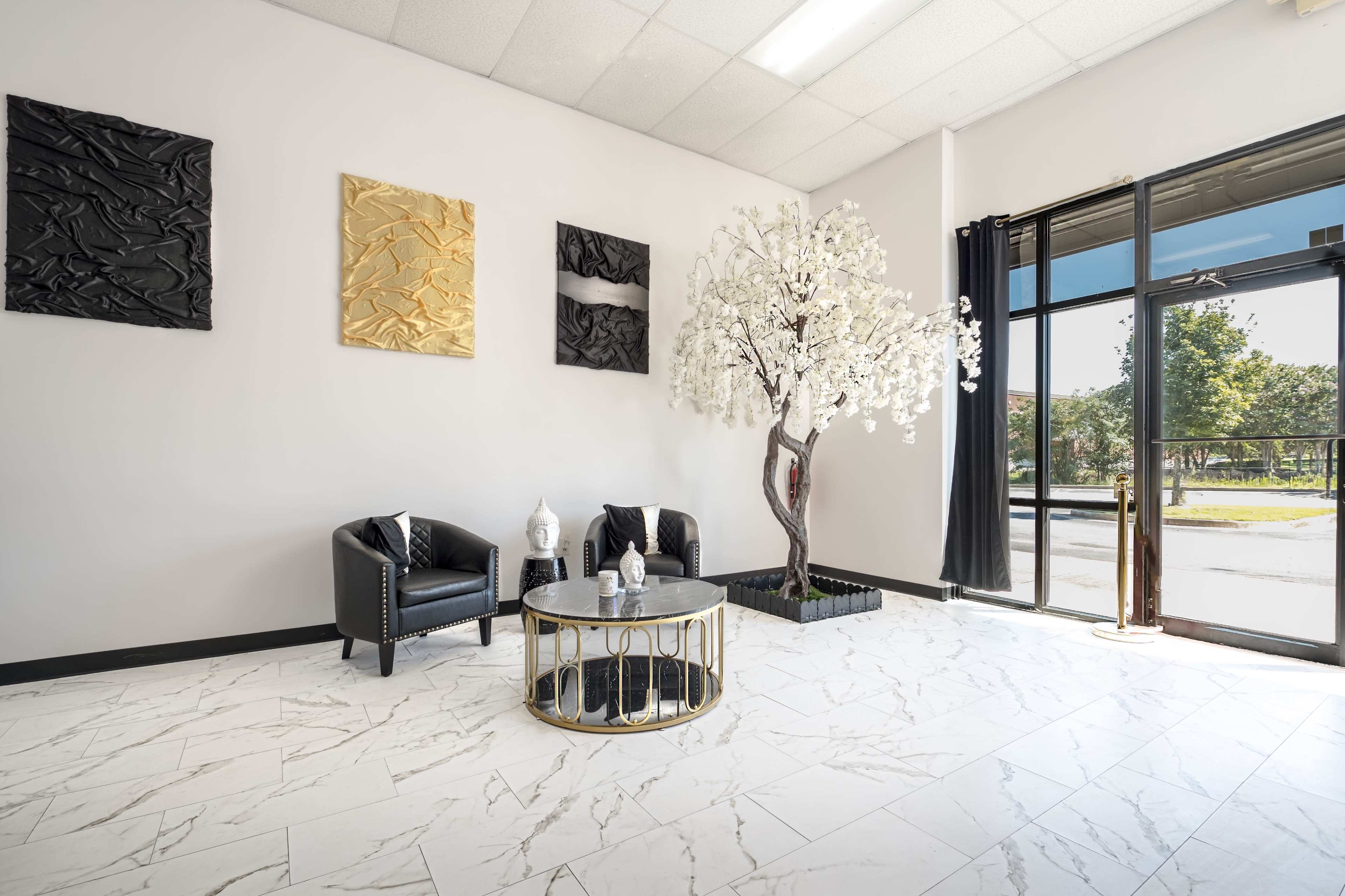 A modern interior features black chairs, a circular coffee table, decorative wall art, and a faux flowering tree near a large glass door.