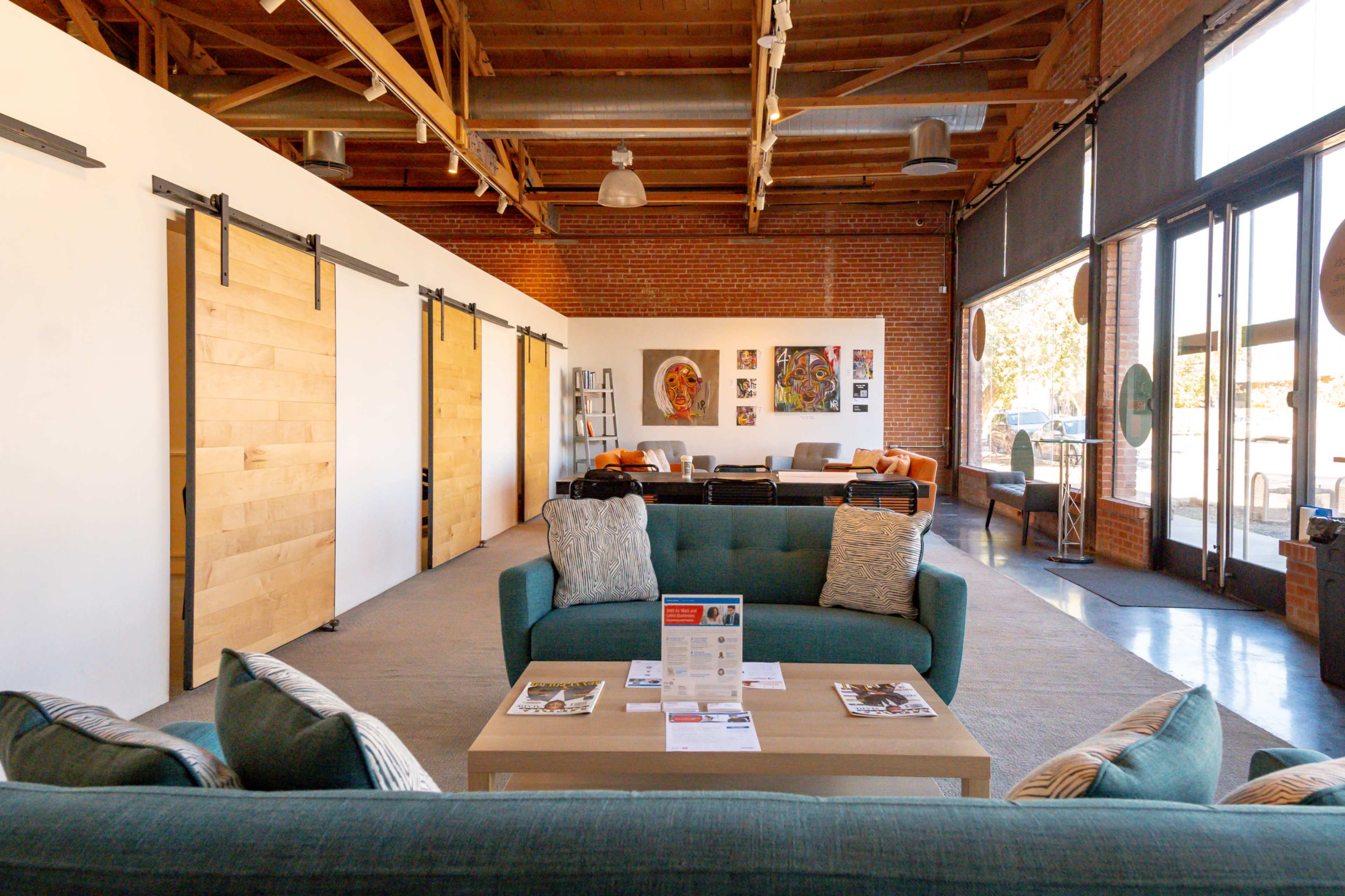 A modern, open-concept interior featuring a seating area with teal sofas, a coffee table, and wooden sliding doors, alongside walls adorned with artwork in a well-lit space.