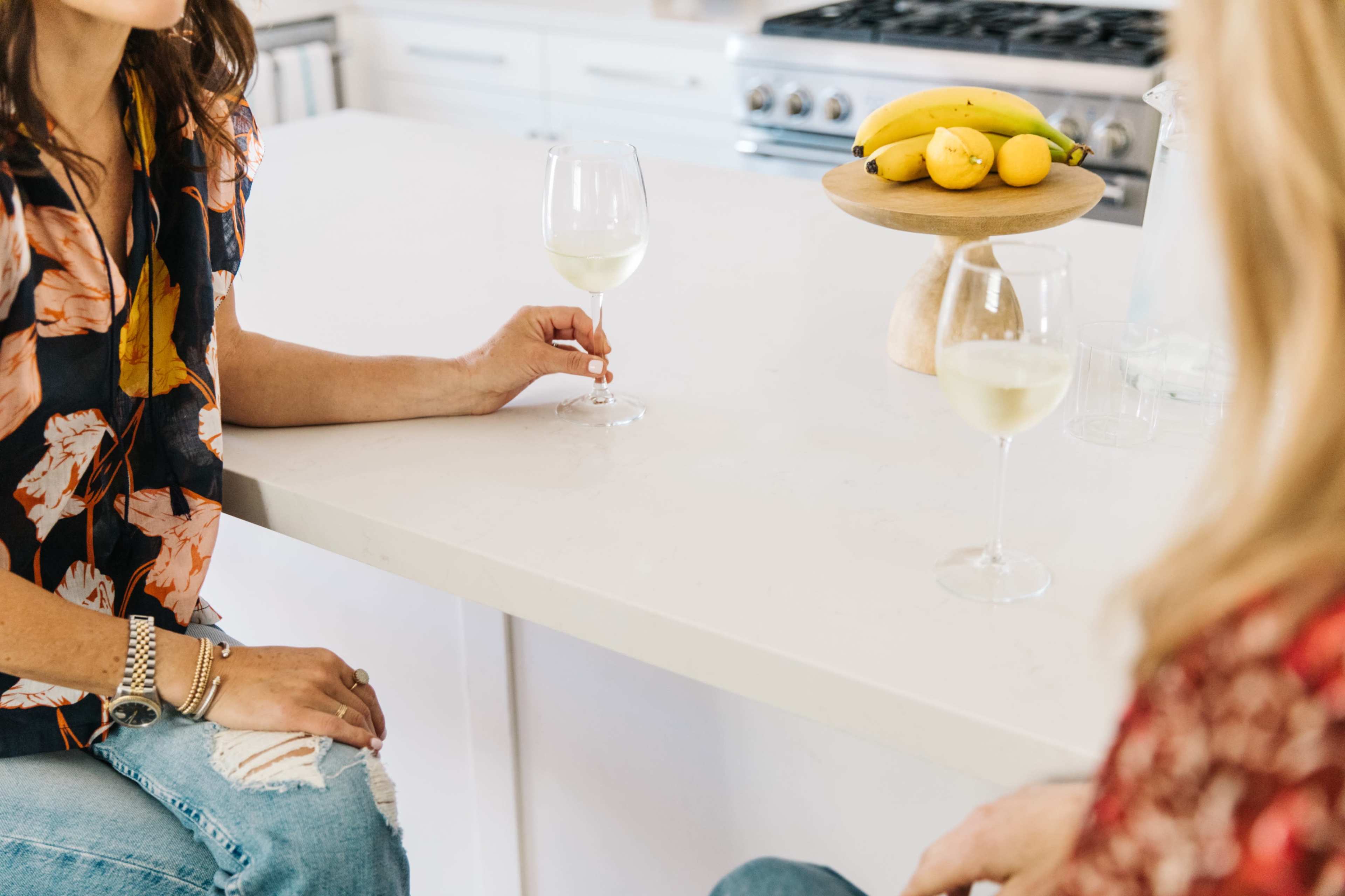 Two women sit at a white kitchen counter with glasses of white wine, while a bowl of bananas and lemons is displayed nearby.