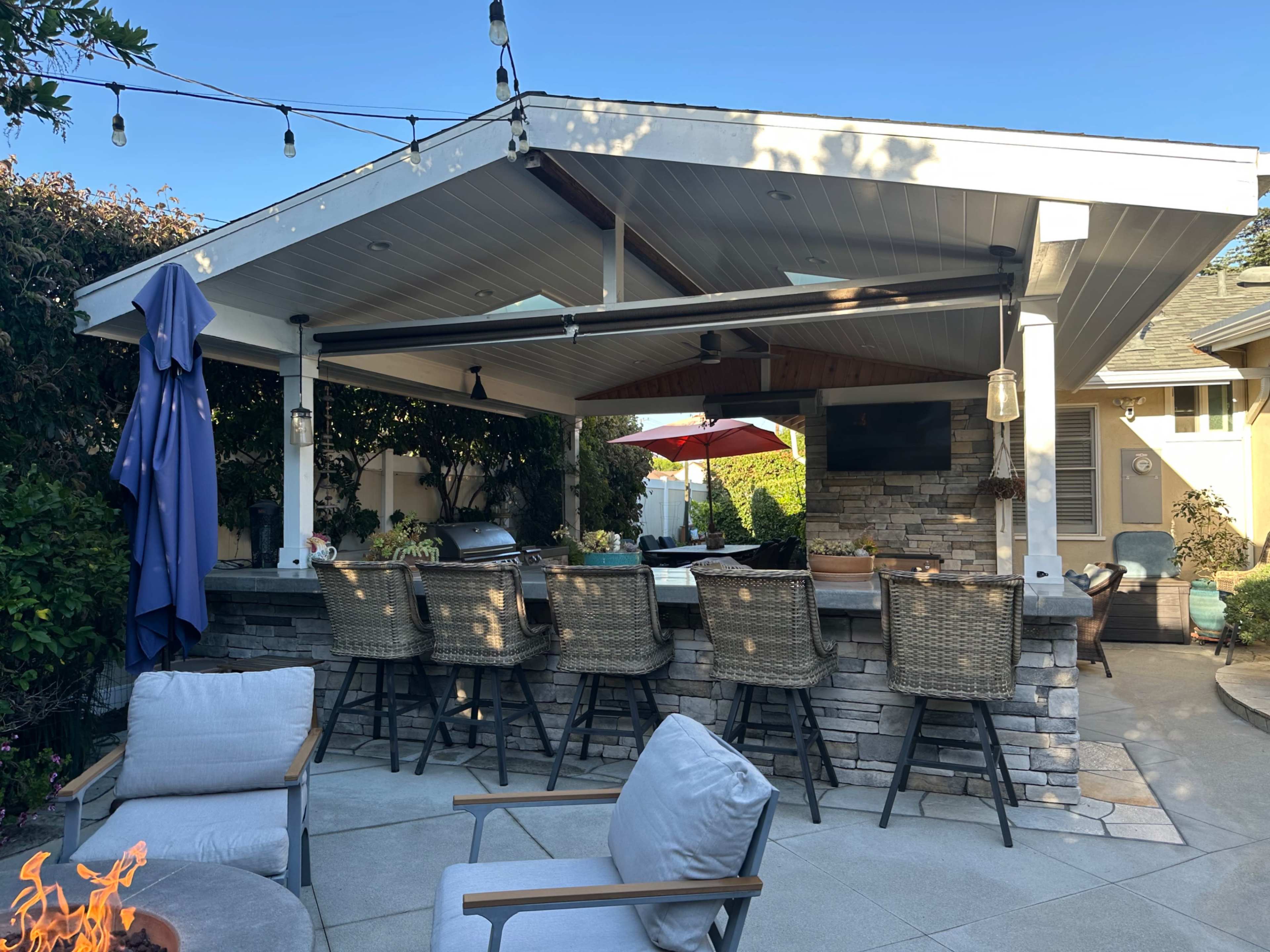 The image shows a covered outdoor kitchen and bar area with high stools, a stone wall, and string lights above, surrounded by greenery.