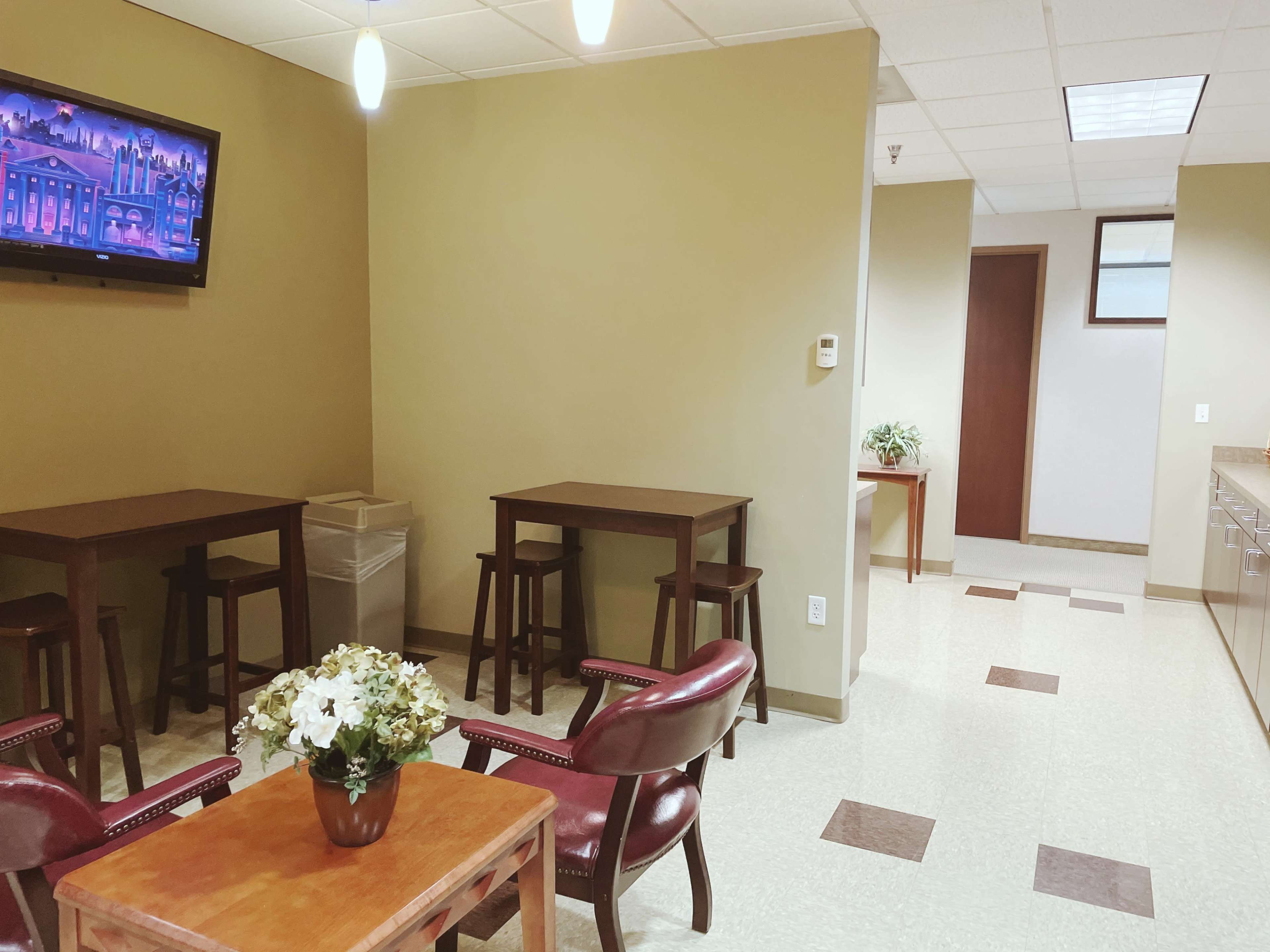 A small waiting area with two wooden tables, two chairs, a television on the wall, and a flower arrangement on a coffee table.
