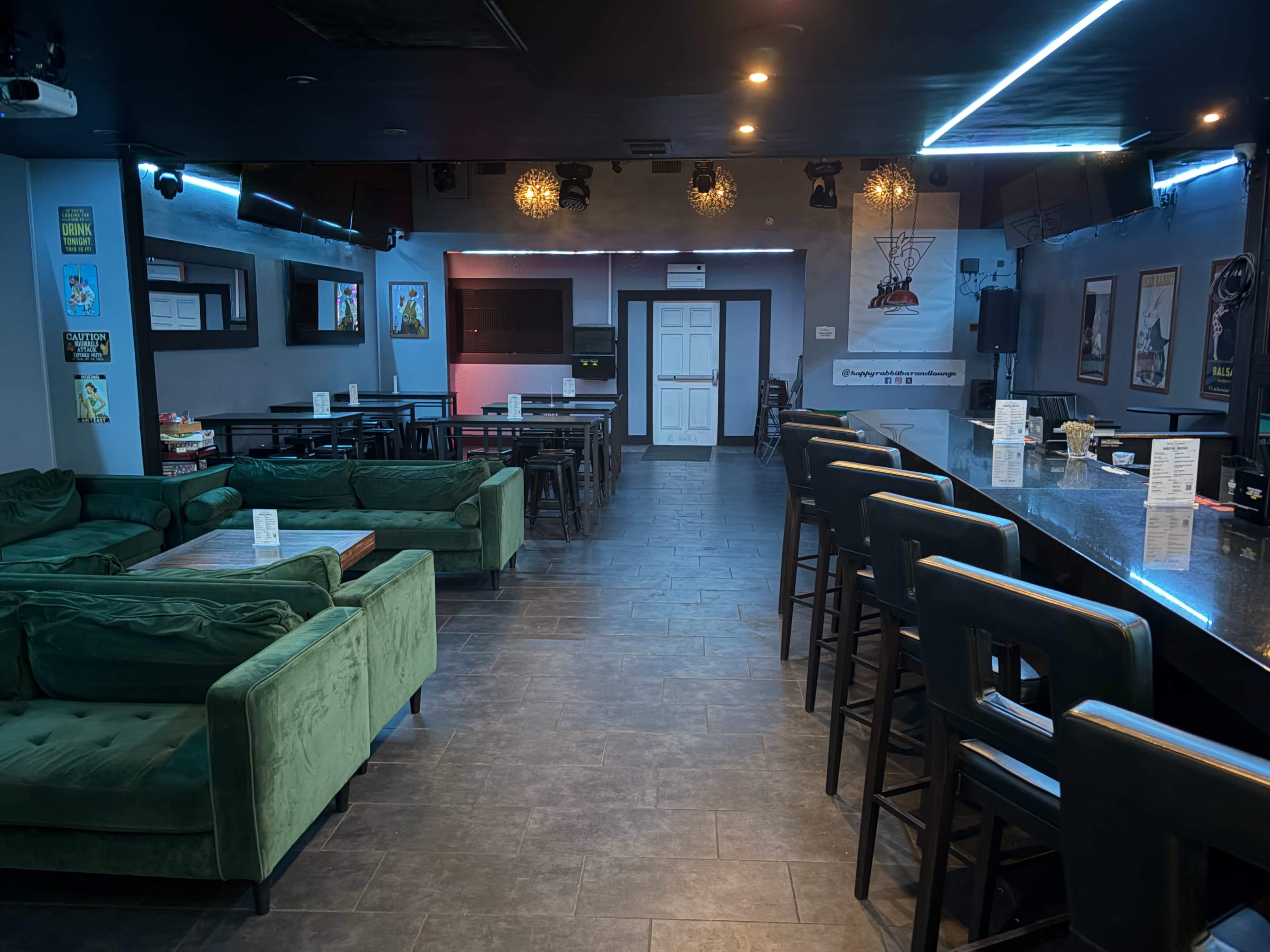 The image shows a modern lounge area featuring green couches, bar stools, and a long counter, with a stage at the far end illuminated by colored lights.
