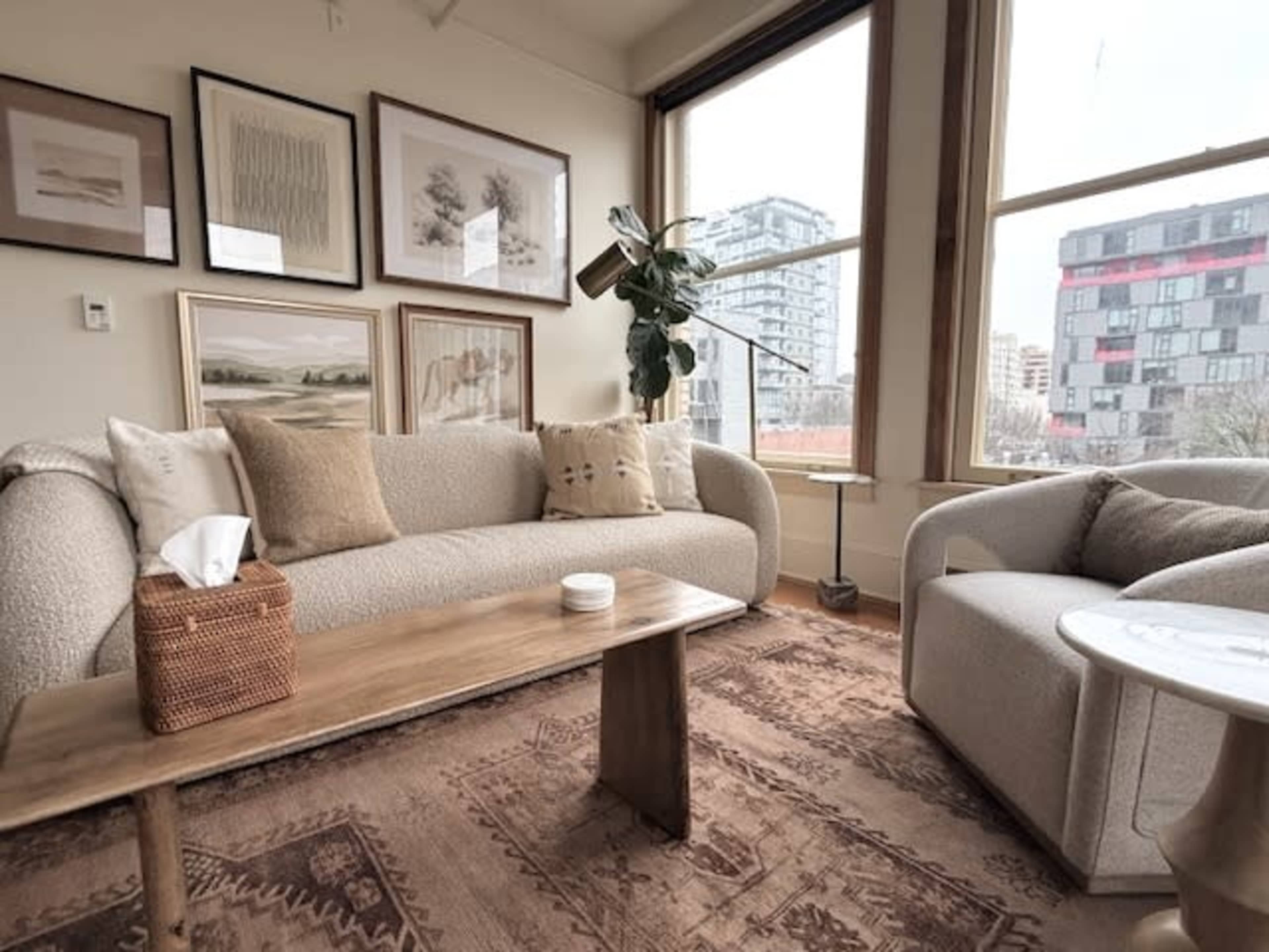 A cozy living room features a light-colored sofa and armchair, a wooden coffee table, and framed artwork on the walls, with large windows providing natural light and a city view.