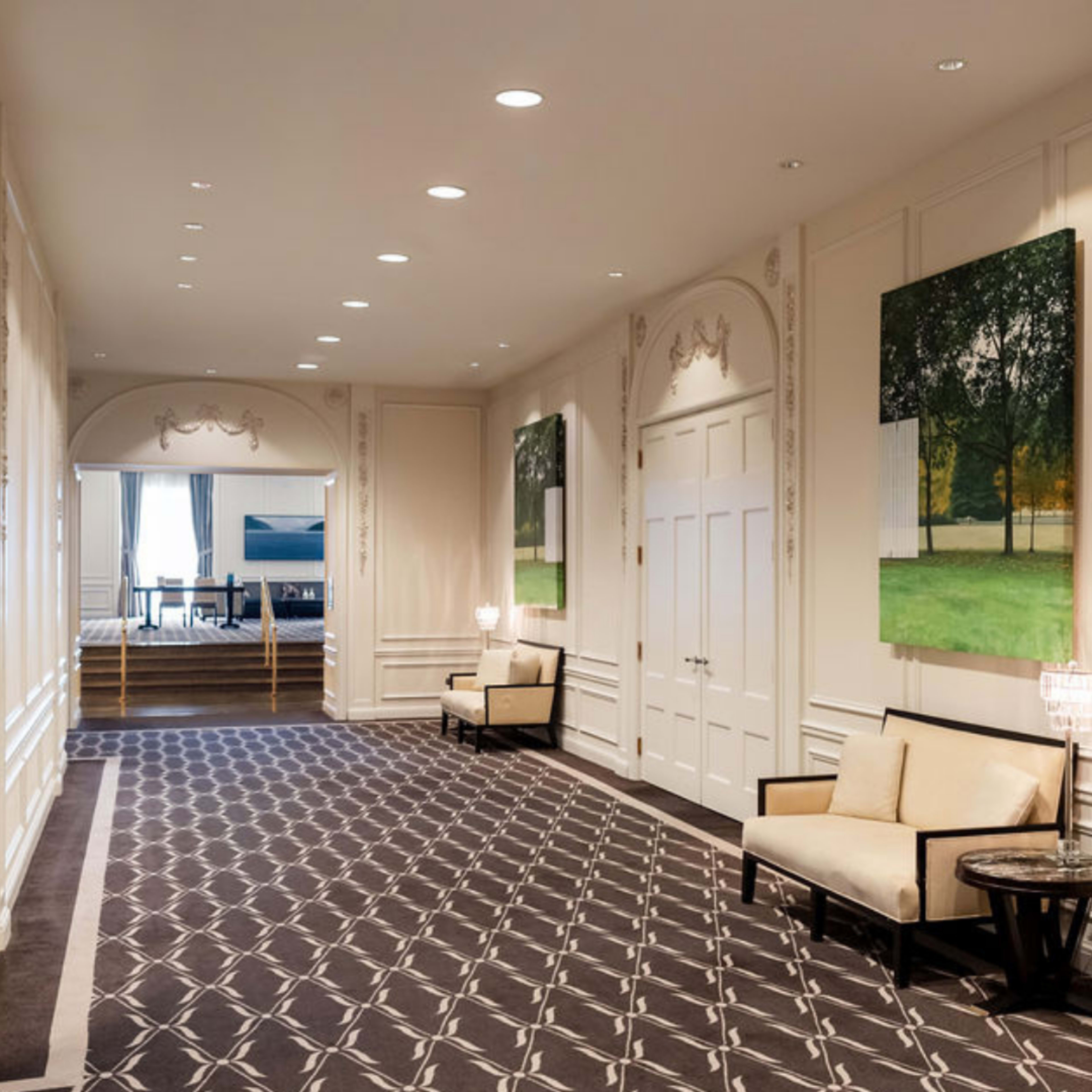 The image depicts a elegant hotel hallway featuring two armchairs, large wall art, and well-lit space with patterned carpet.