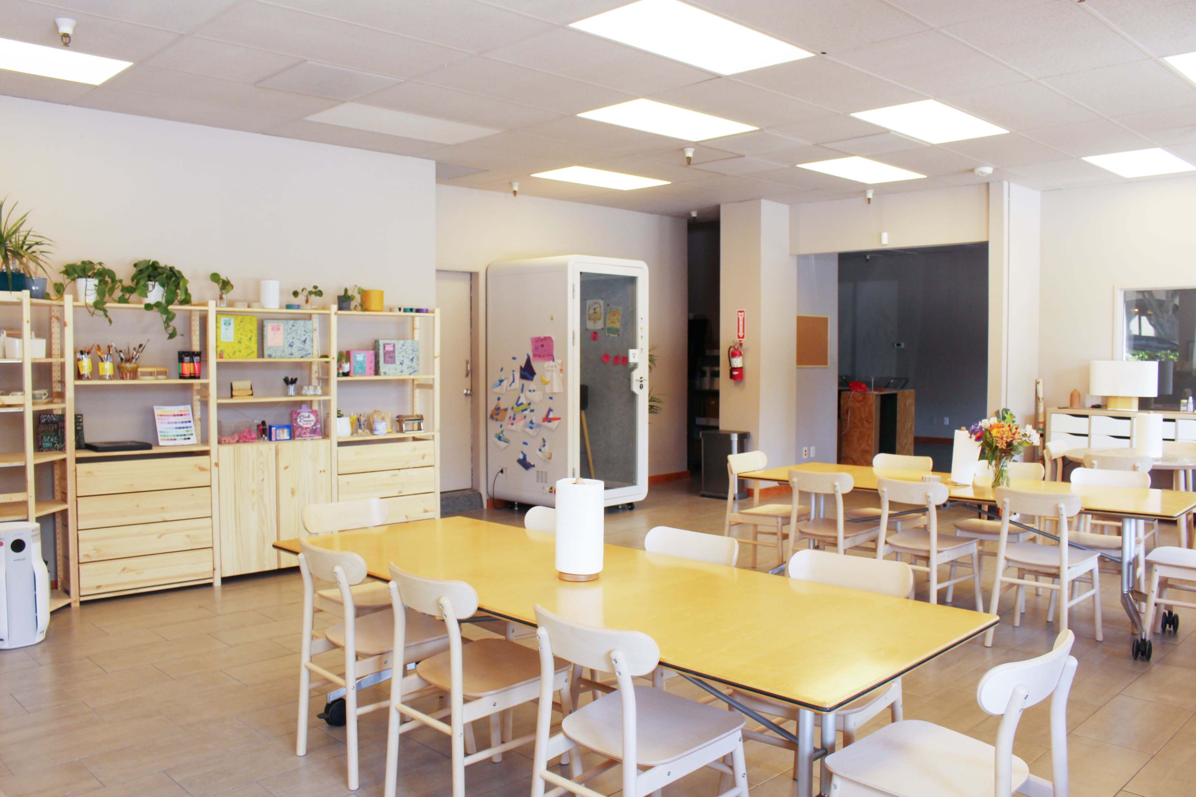 The image shows a spacious, well-lit room with light-colored furniture, wooden tables, and chairs, along with shelves containing various items against the walls.
