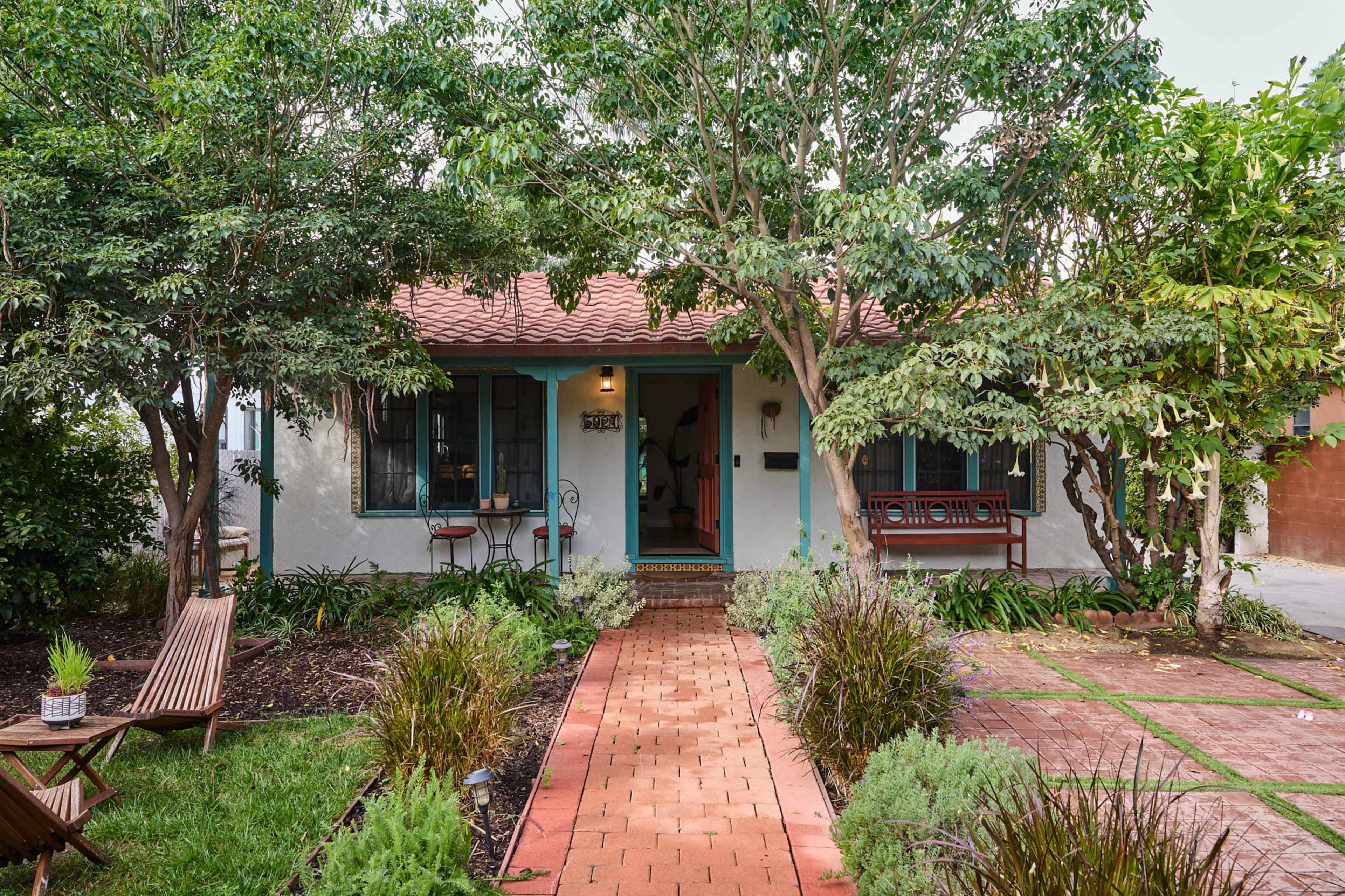A small, single-story house with a red tile roof is surrounded by lush greenery and a brick pathway leading to the front door.