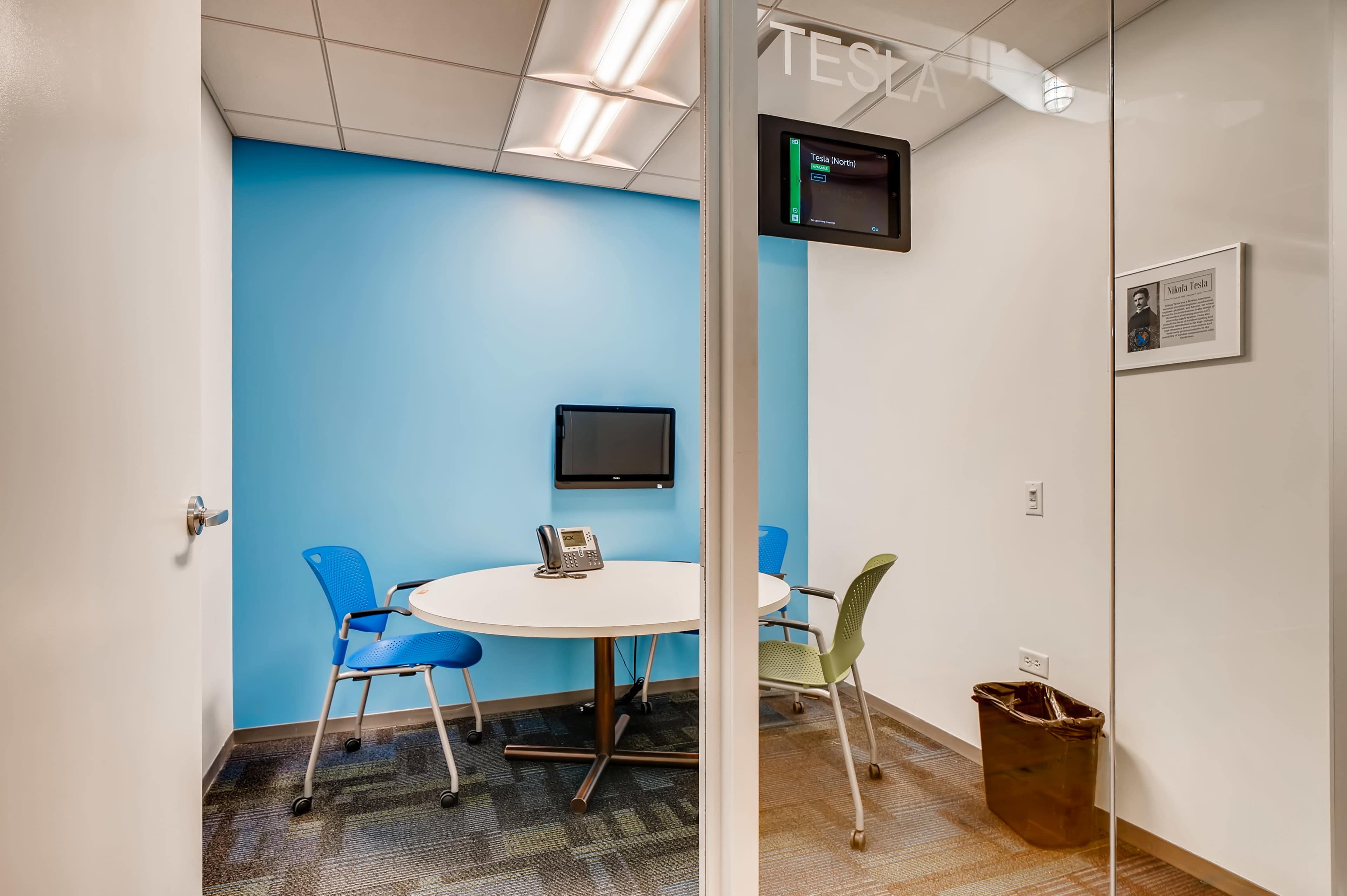 The image shows a small conference room with a round table, two chairs, a wall-mounted monitor, and a telephone, featuring a blue accent wall.