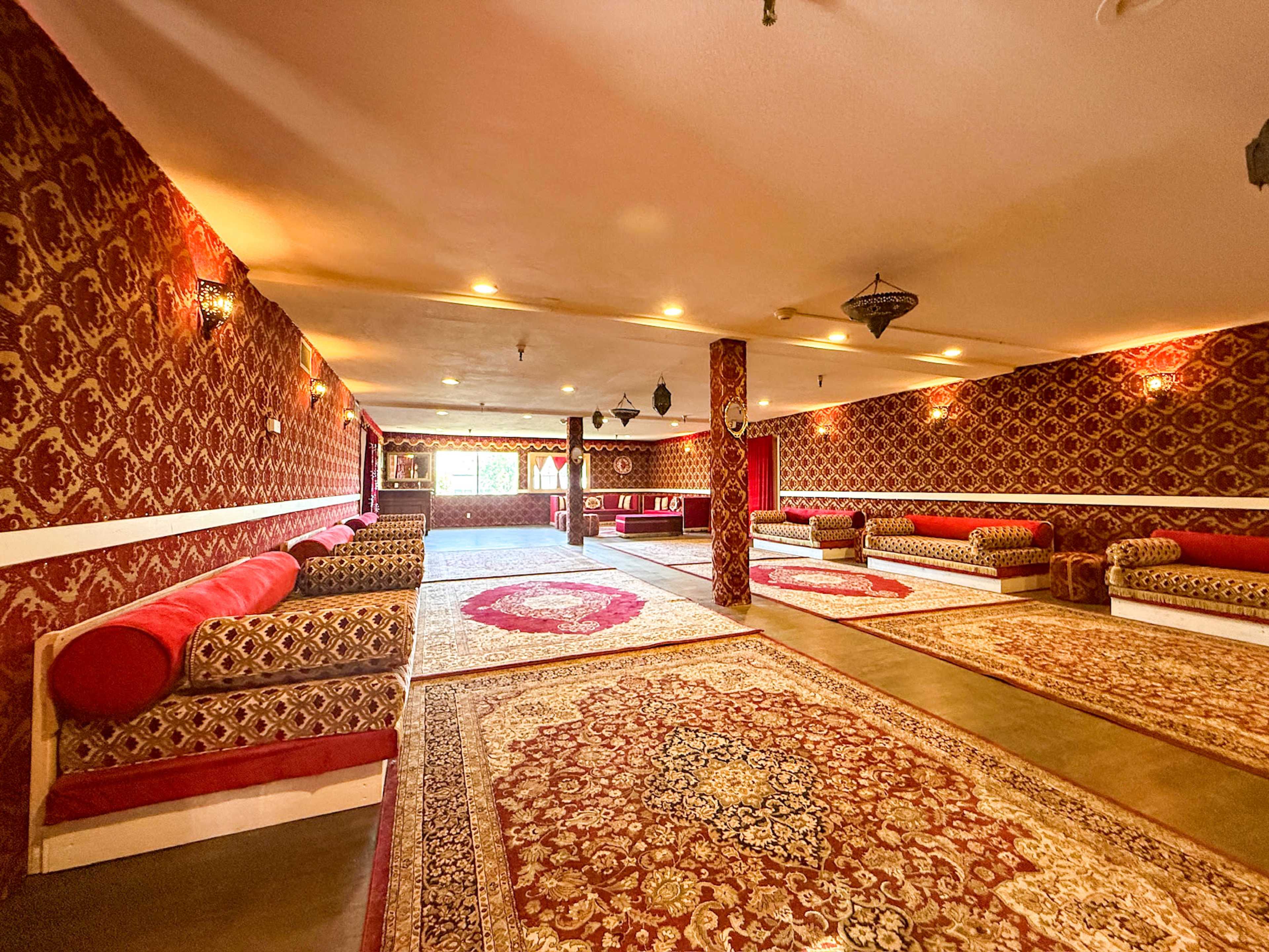The image shows a spacious room decorated with intricately patterned red wallpaper, multiple area rugs, and seating along the walls.