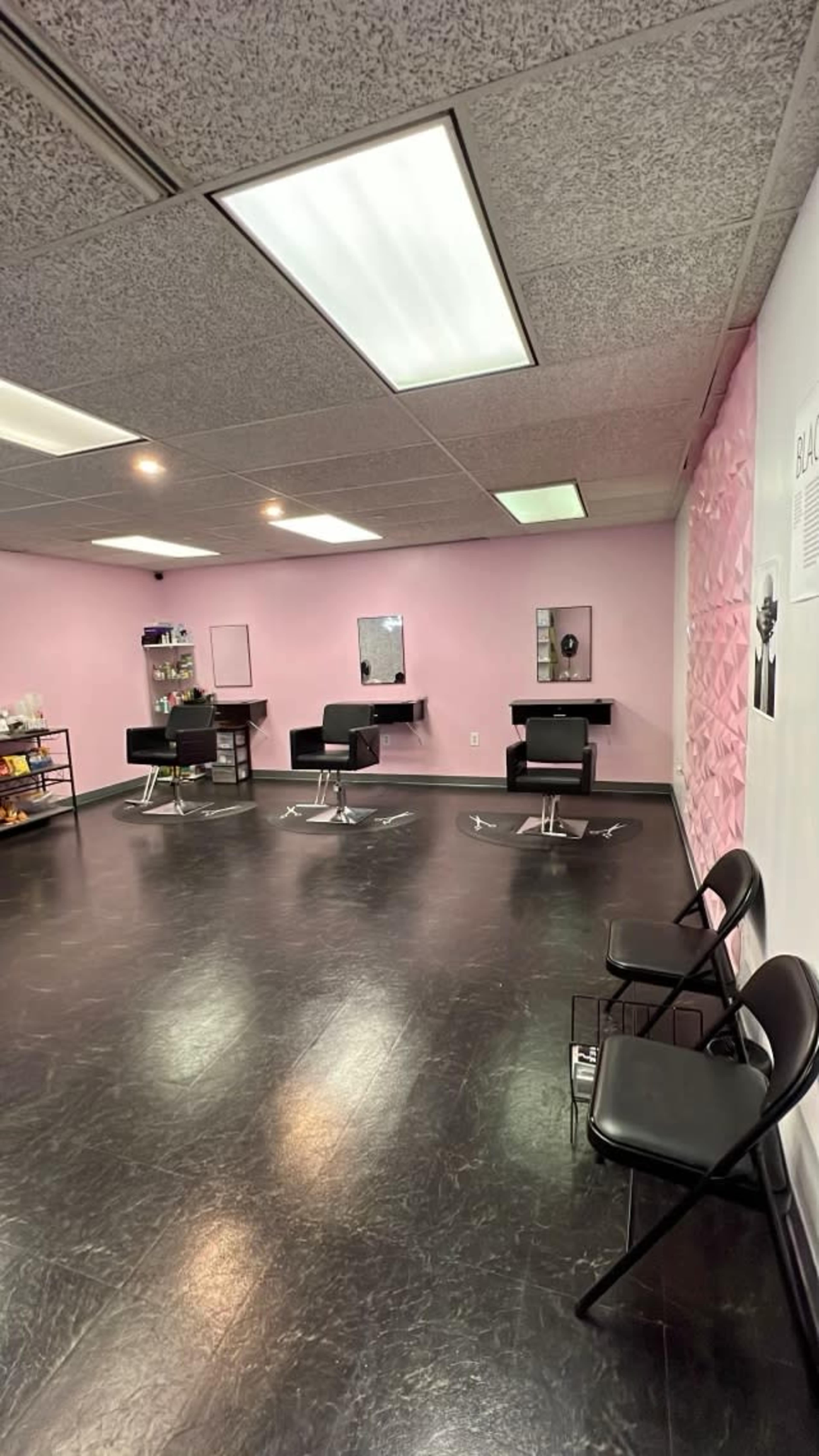 A hair salon interior with black flooring, three black styling chairs, and a pink wall with mirrors.