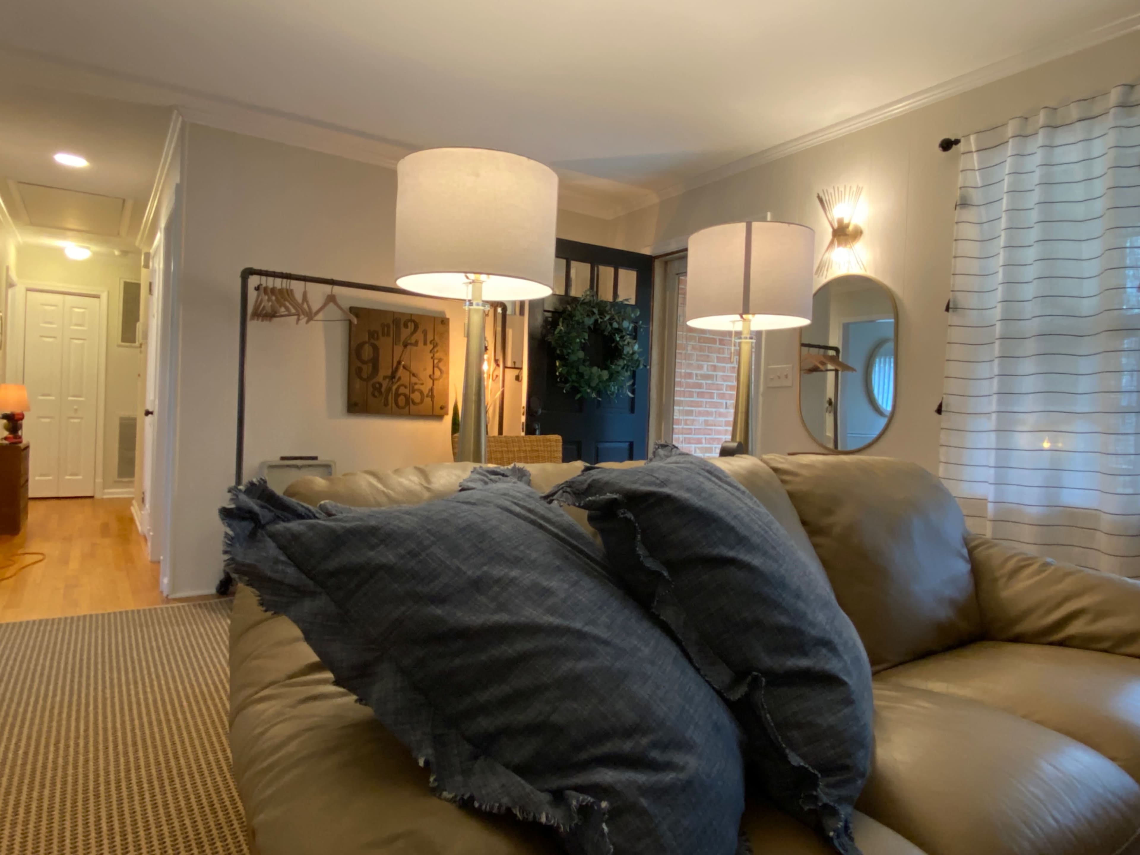 A living room features a beige sofa with blue pillows, two floor lamps, a round mirror, and a doorway leading to a hallway.
