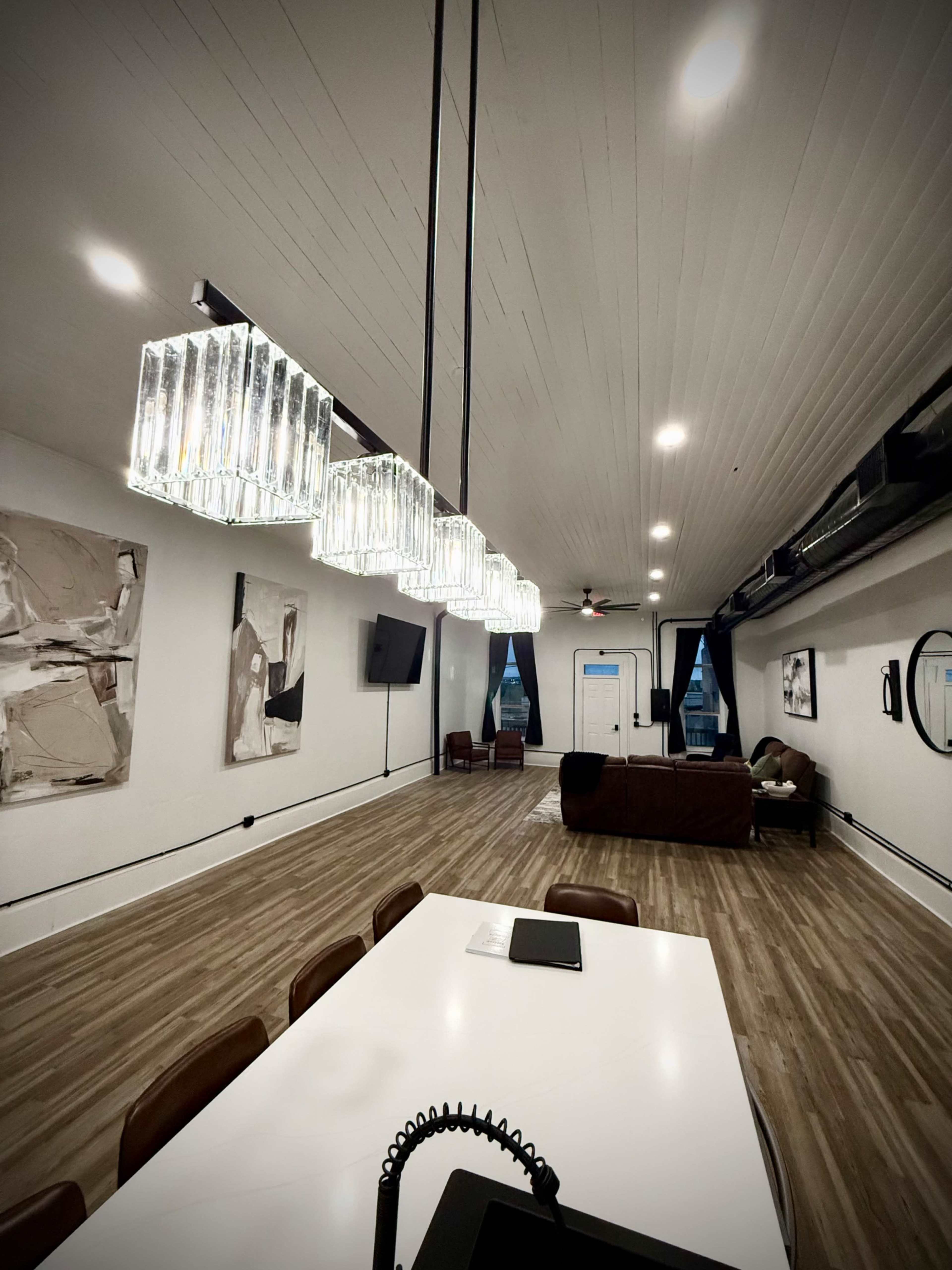 The image shows a modern interior space featuring a long dining table, a living area with a dark sofa, and pendant lighting hanging from the ceiling.