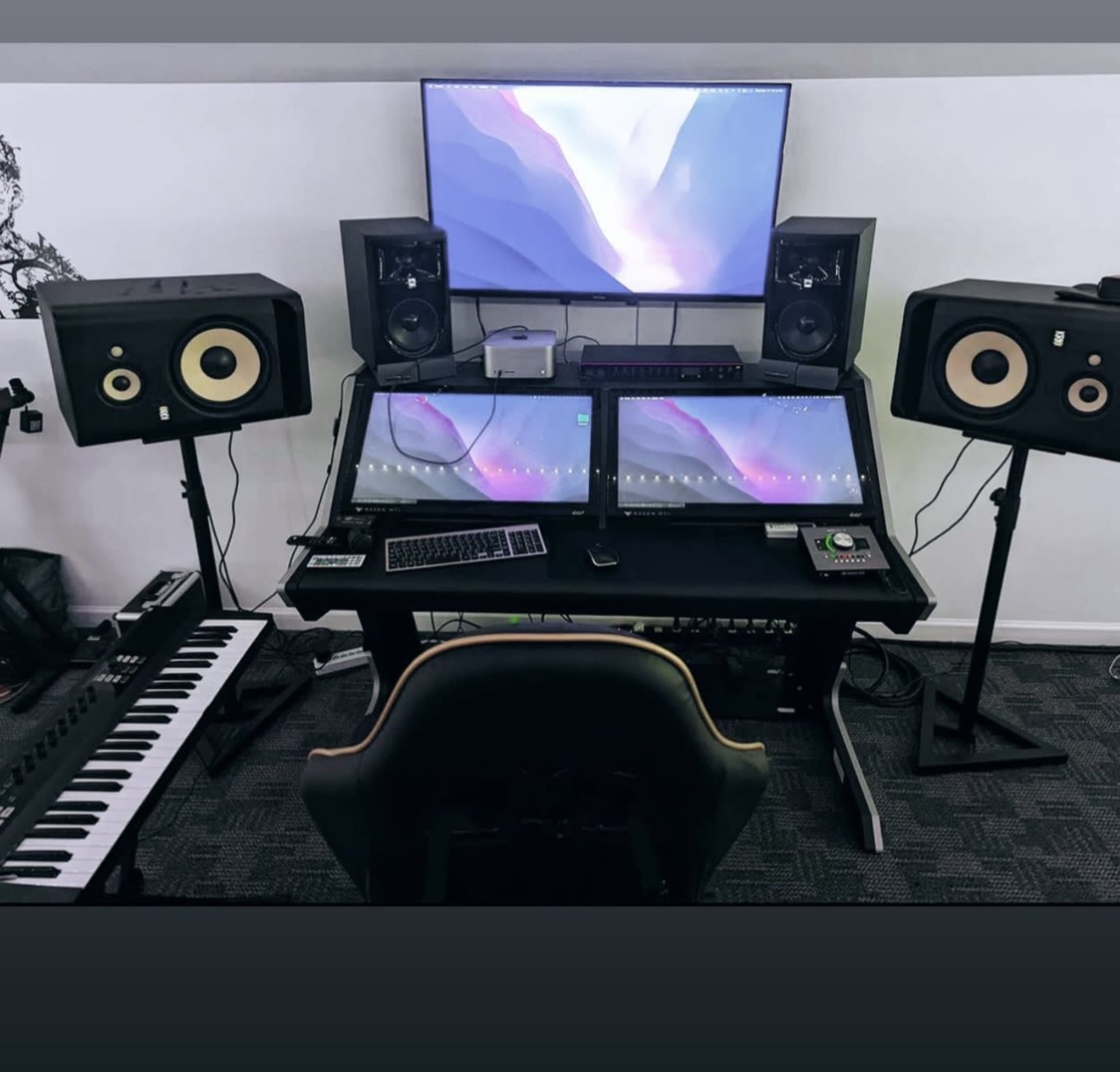 A music production setup with dual monitors, studio speakers, a keyboard, and a control surface on a desk.