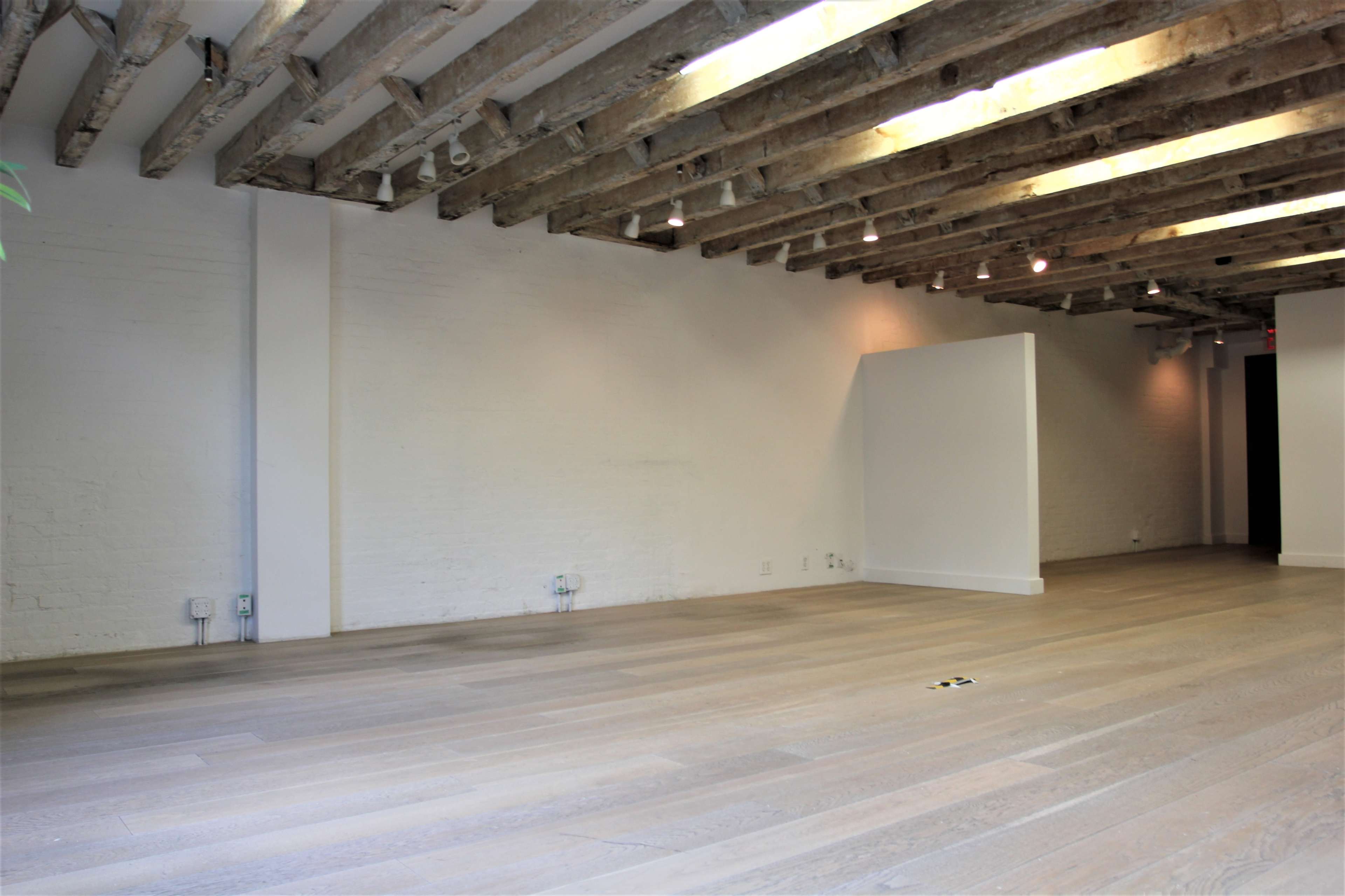 The image shows an empty interior space with wooden beams on the ceiling, light-colored wooden flooring, and white walls.