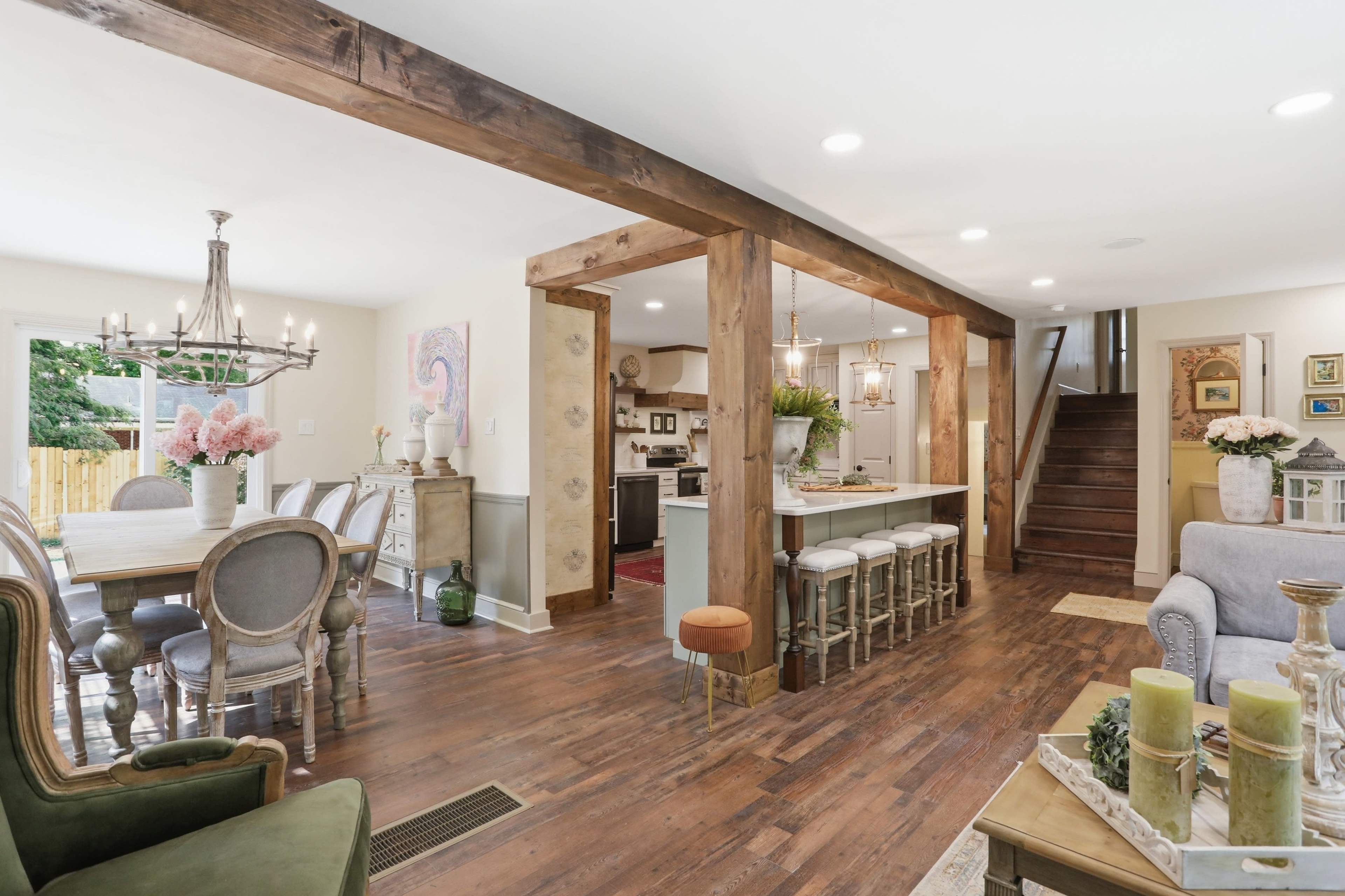 The image shows an open-concept dining and kitchen area with wooden beams, a long dining table, and a staircase in the background.
