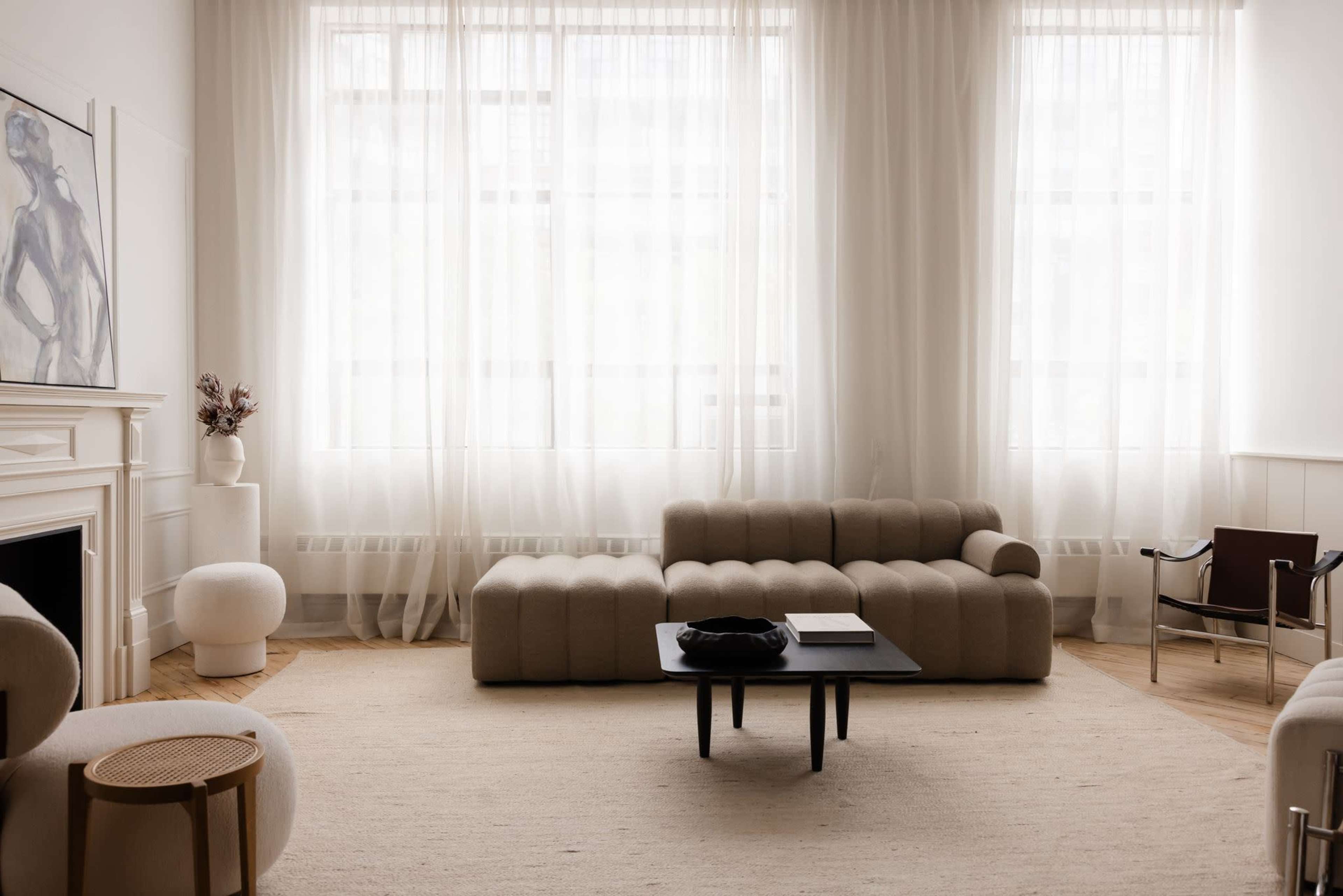 The image shows a minimalist living room with large windows covered by sheer curtains, a beige sectional sofa, a black coffee table, and a small round side table.
