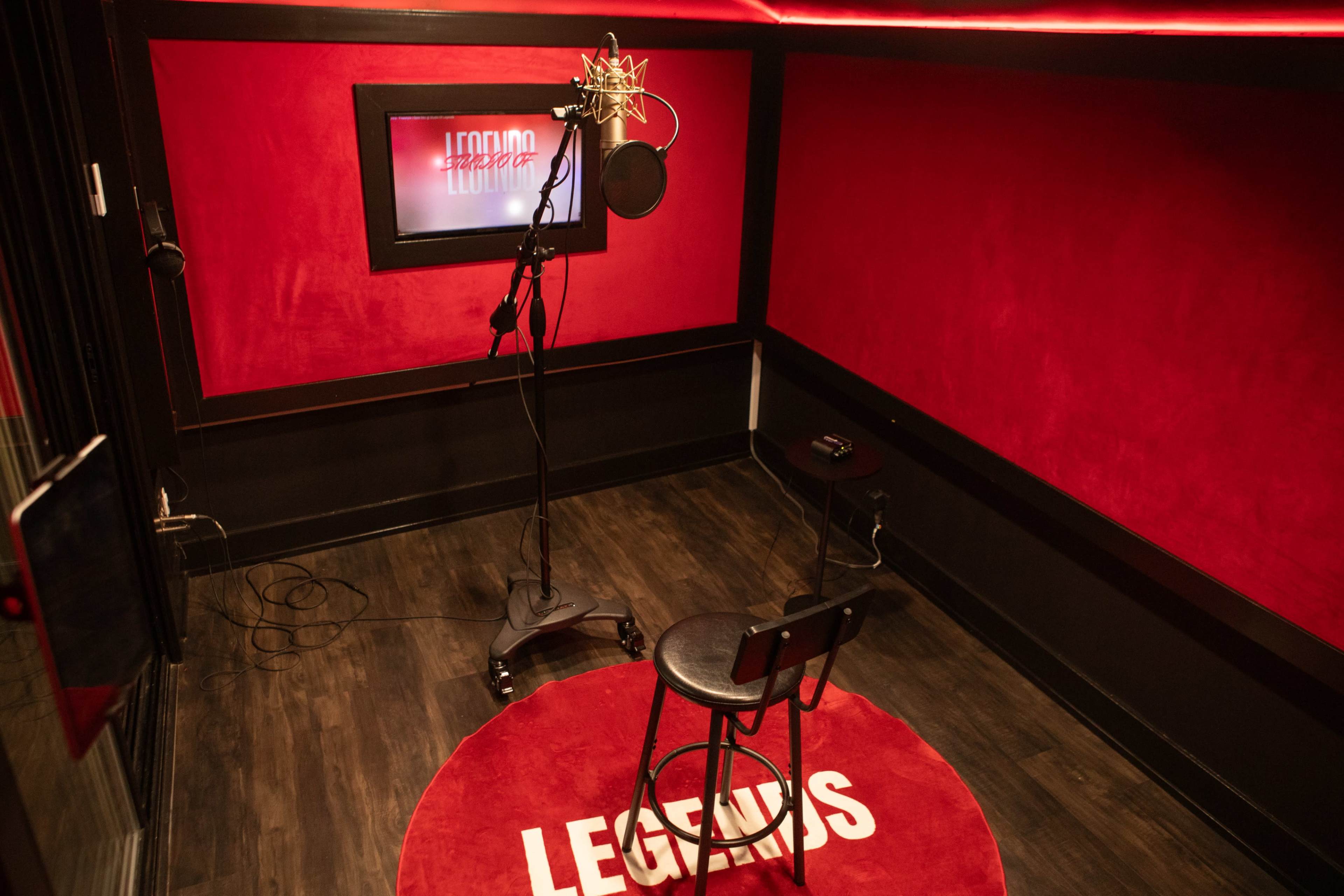 The image shows a small soundproof recording studio with red walls, a microphone on a stand, and a black stool on a round red rug that says "LEGENDS."